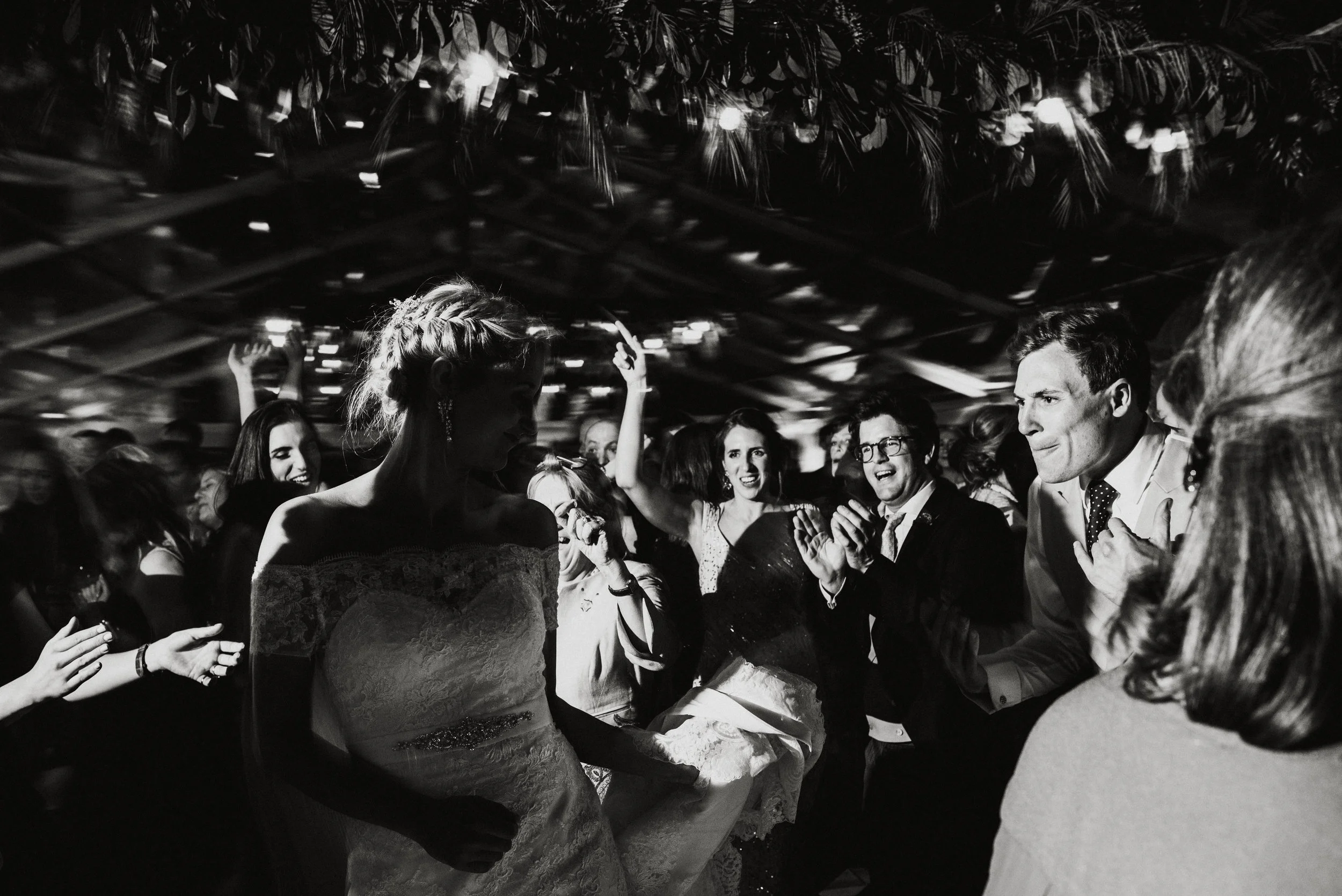 People dancing and celebrating at a wedding reception, some smiling and clapping, in a decorated indoor venue.