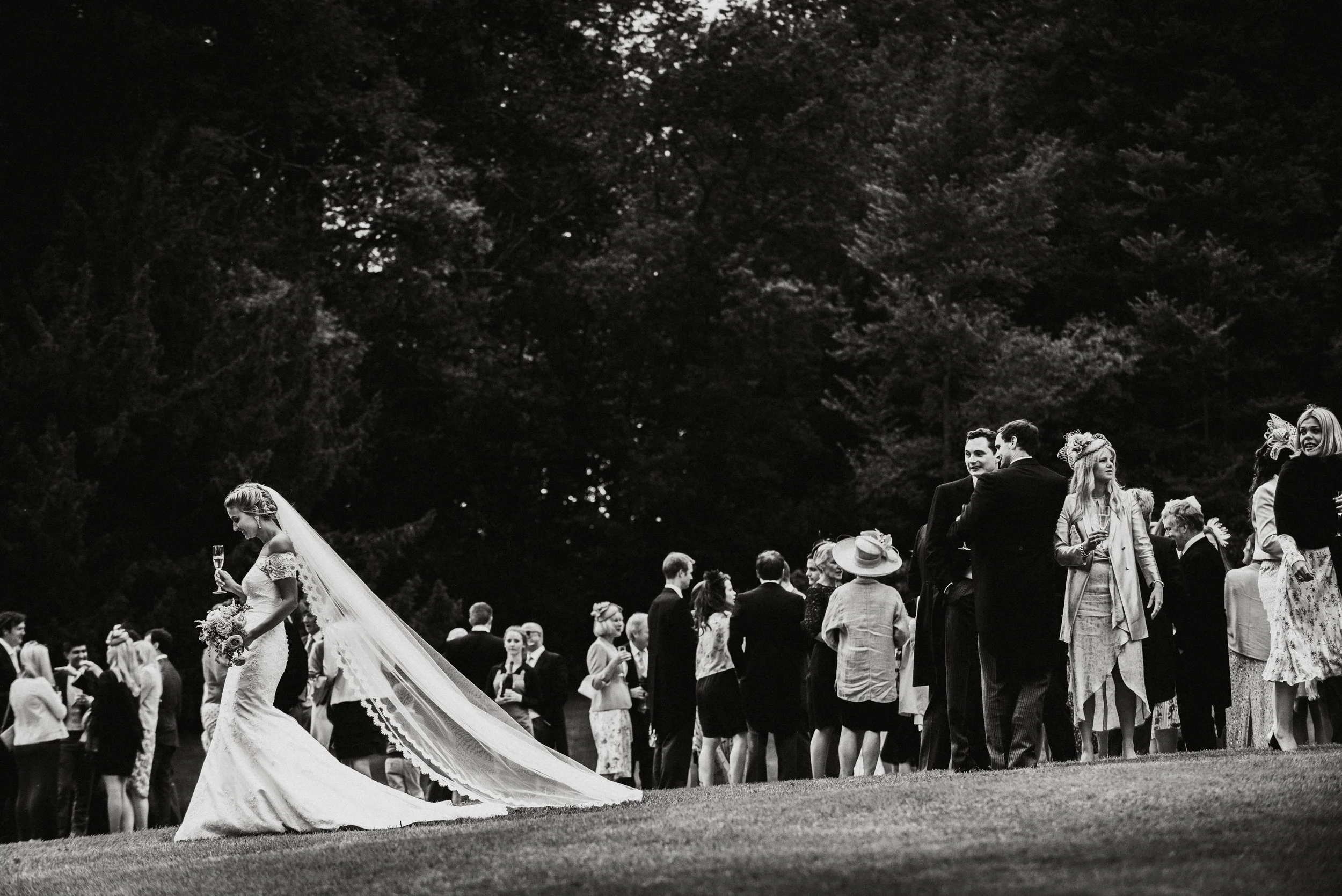 A bride holding a glass of champagne and a bouquet, with a long veil trailing behind her, standing alone on a grassy area at a wedding reception, while groups of guests in formal attire mingle and converse in the background, with a backdrop of trees,