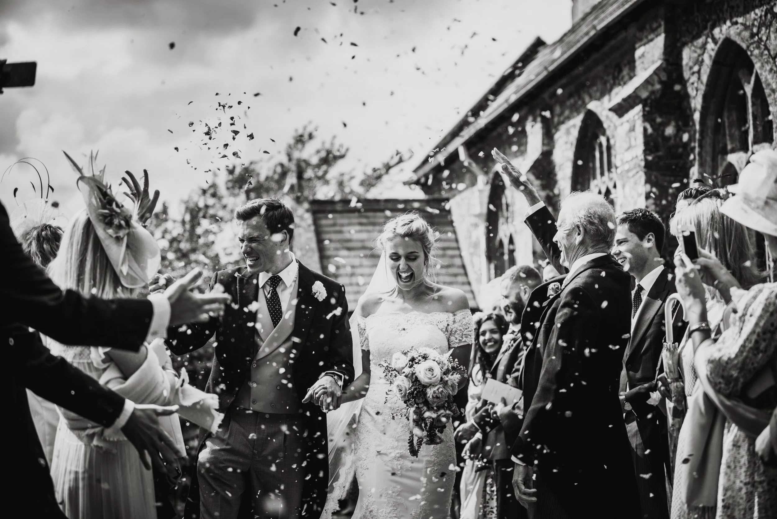Wedding photo in black and white of confetti celebrations with the bride and groom smiling amidst guests throwing confetti outside a rustic building in Cornwall.