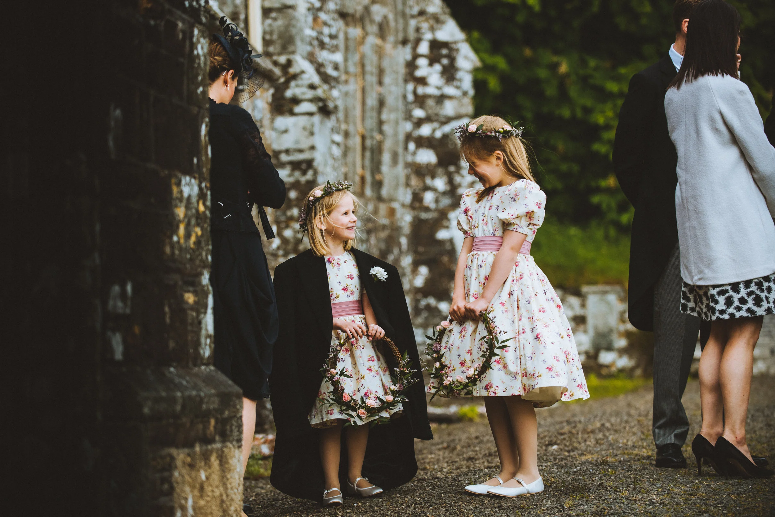 Isles of scilly photography. Two young girls in floral dresses and floral crowns holding flower wreaths, smiling and talking outdoors near stone ruins, with adults standing nearby.