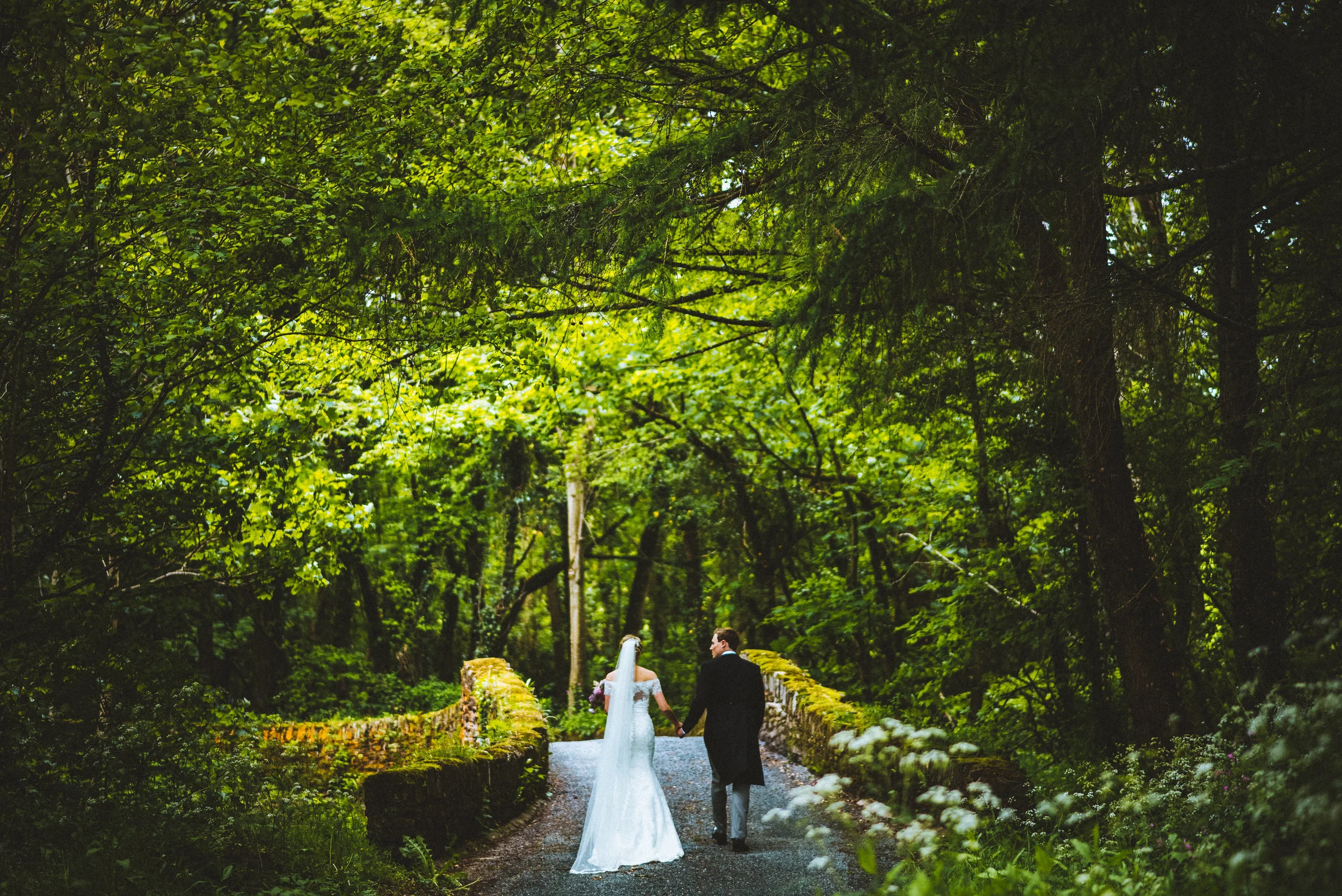 Wedding photo of A bride and groom walking hand in hand on a forest trail surrounded by lush green trees and foliage.