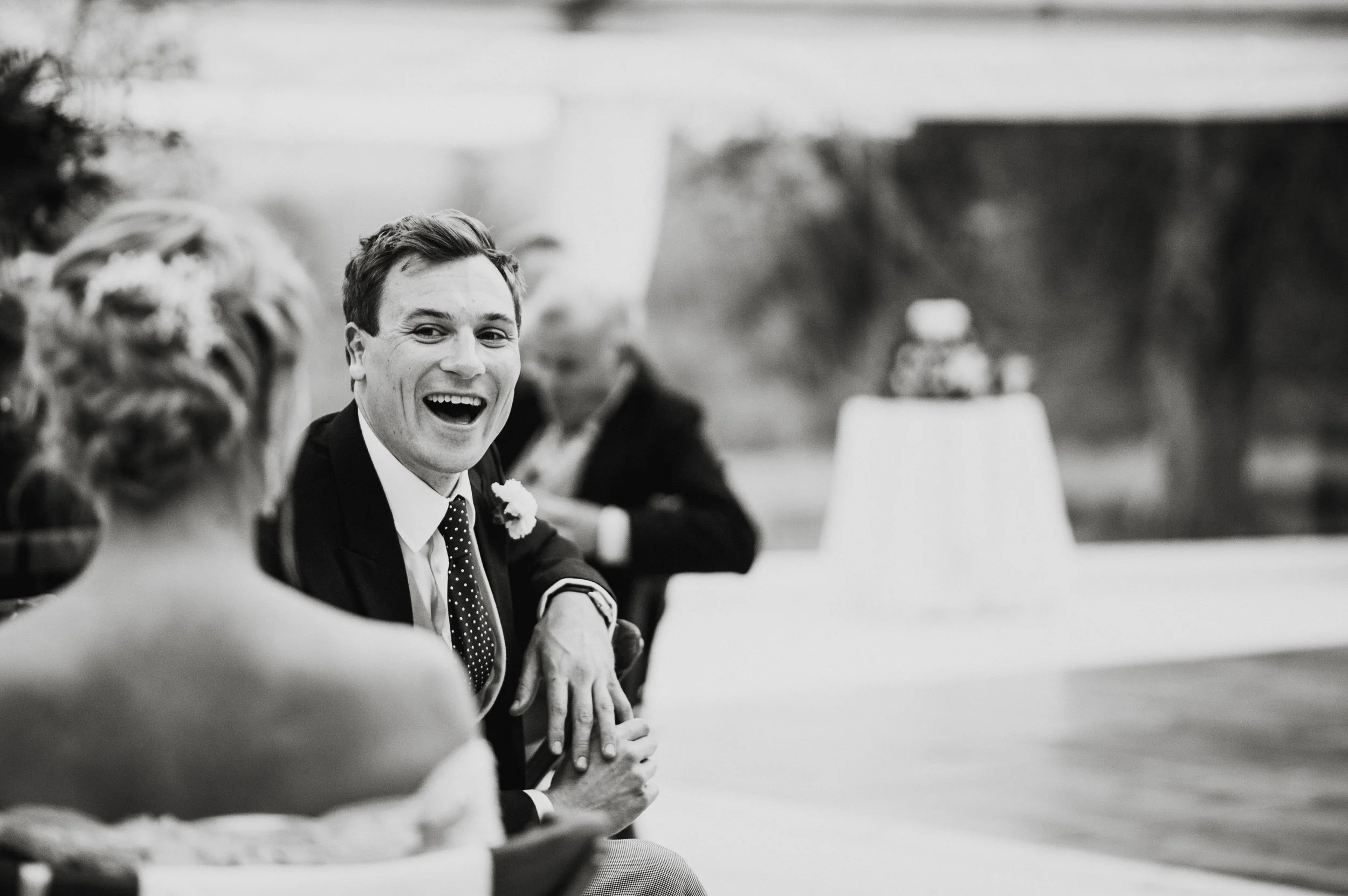 A smiling man in a suit and polka dot tie at a wedding reception, sitting at a table with a woman in a strapless dress and a bun hairstyle.