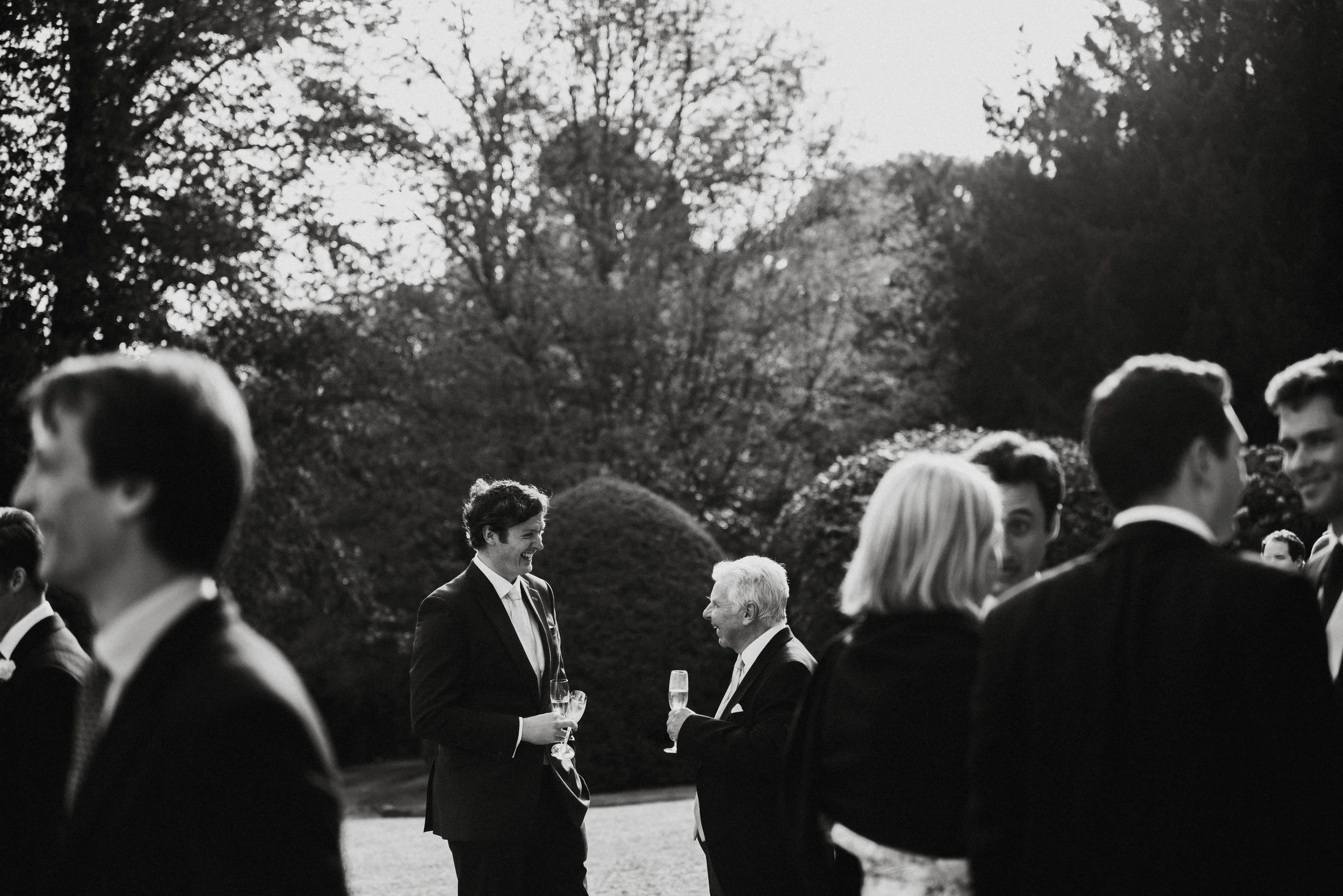 People in formal attire socializing outdoors in a garden, holding glasses of champagne.