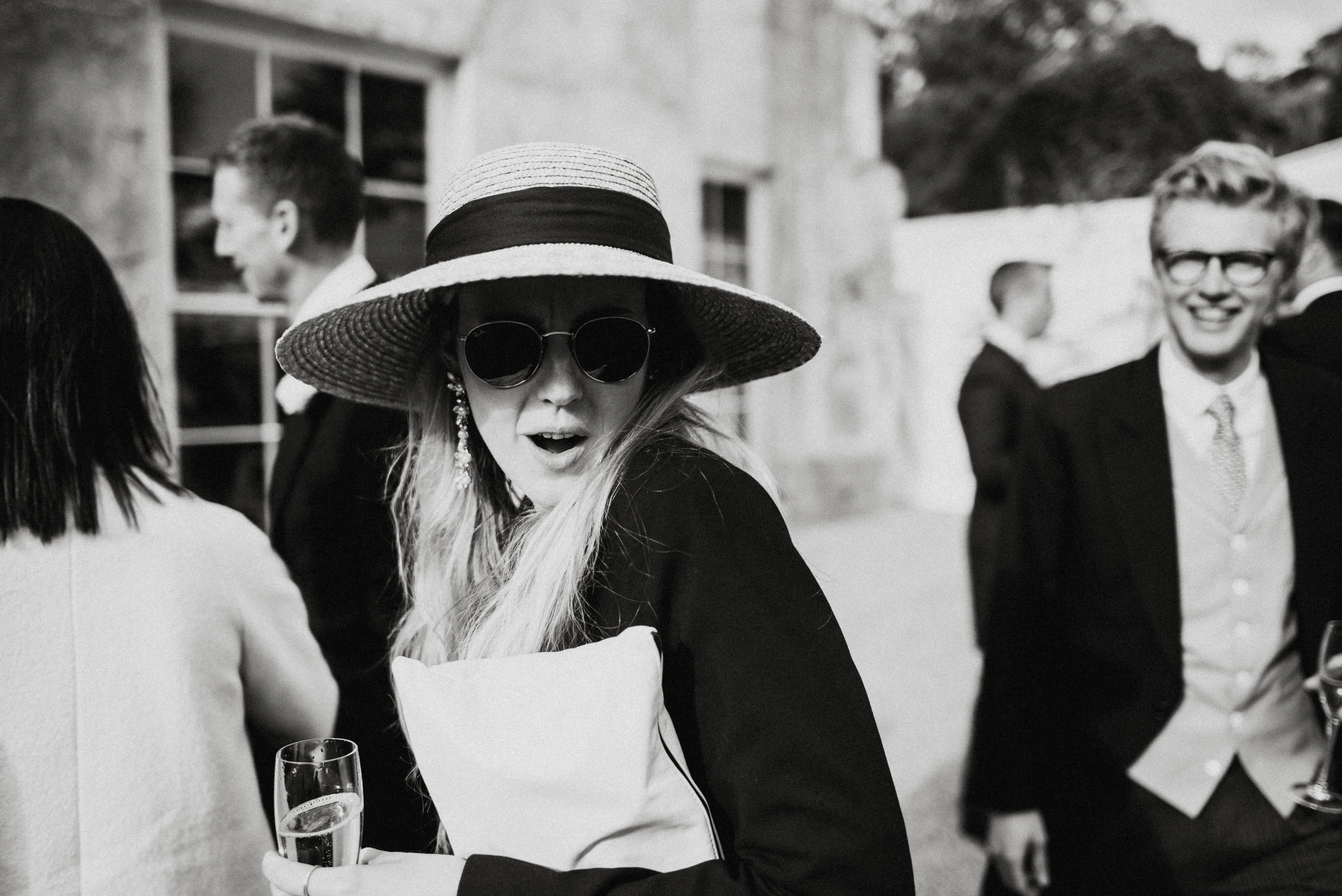 A woman wearing large sunglasses, a wide-brimmed hat, and earrings, holding a champagne flute at a social gathering.