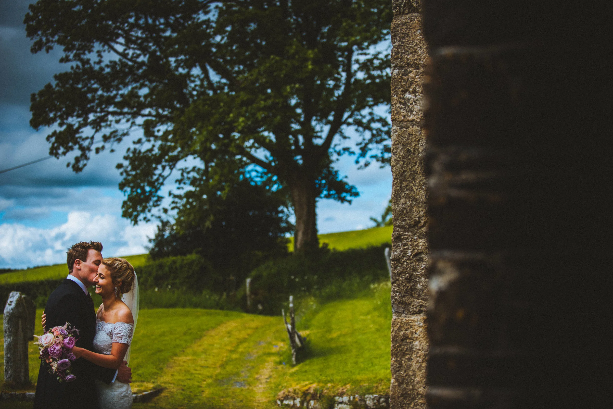 A happy bride and groom embracing outdoors during a wedding, with green hills, trees, a cloudy sky, and a stone wall in the background - Captured by Cornwall based wedding photographer Mark Shaw