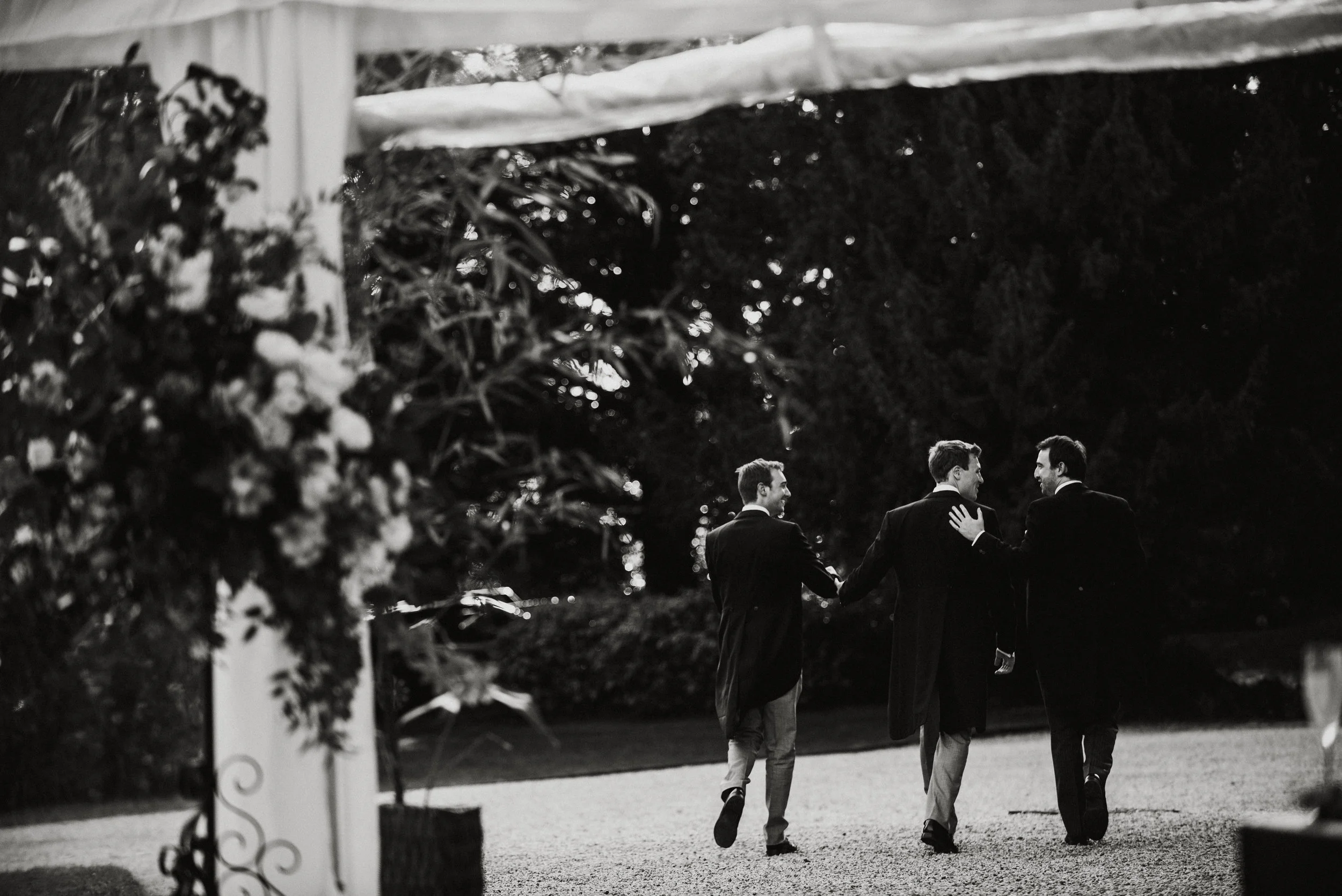Three men dressed in formal suits walking together outdoors at night, with floral decorations visible in the foreground.