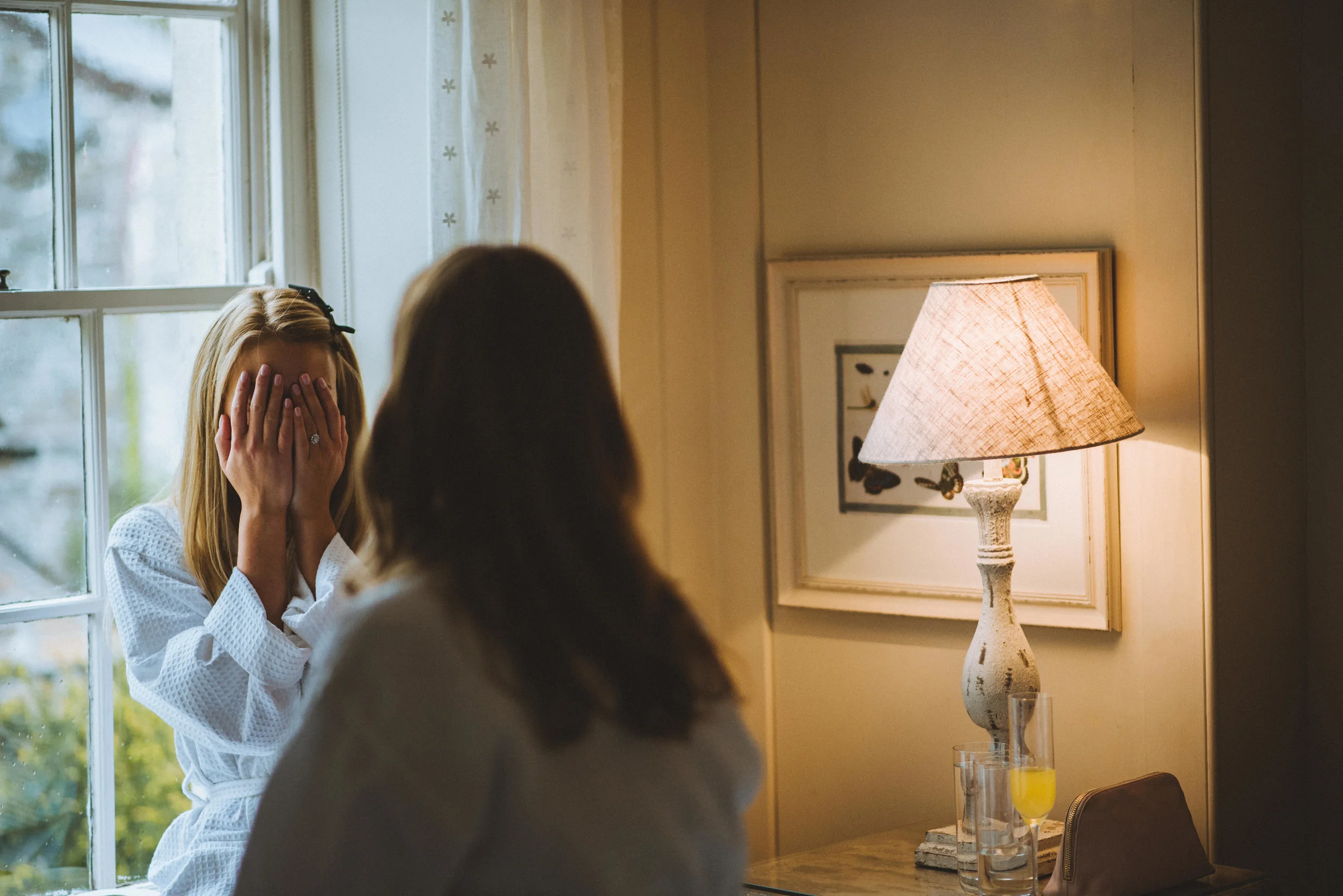 Two women in a cozy room, one covering her face with her hands and the other facing away, near a window with natural light, a table lamp, a framed picture, and drinks on a wooden table.