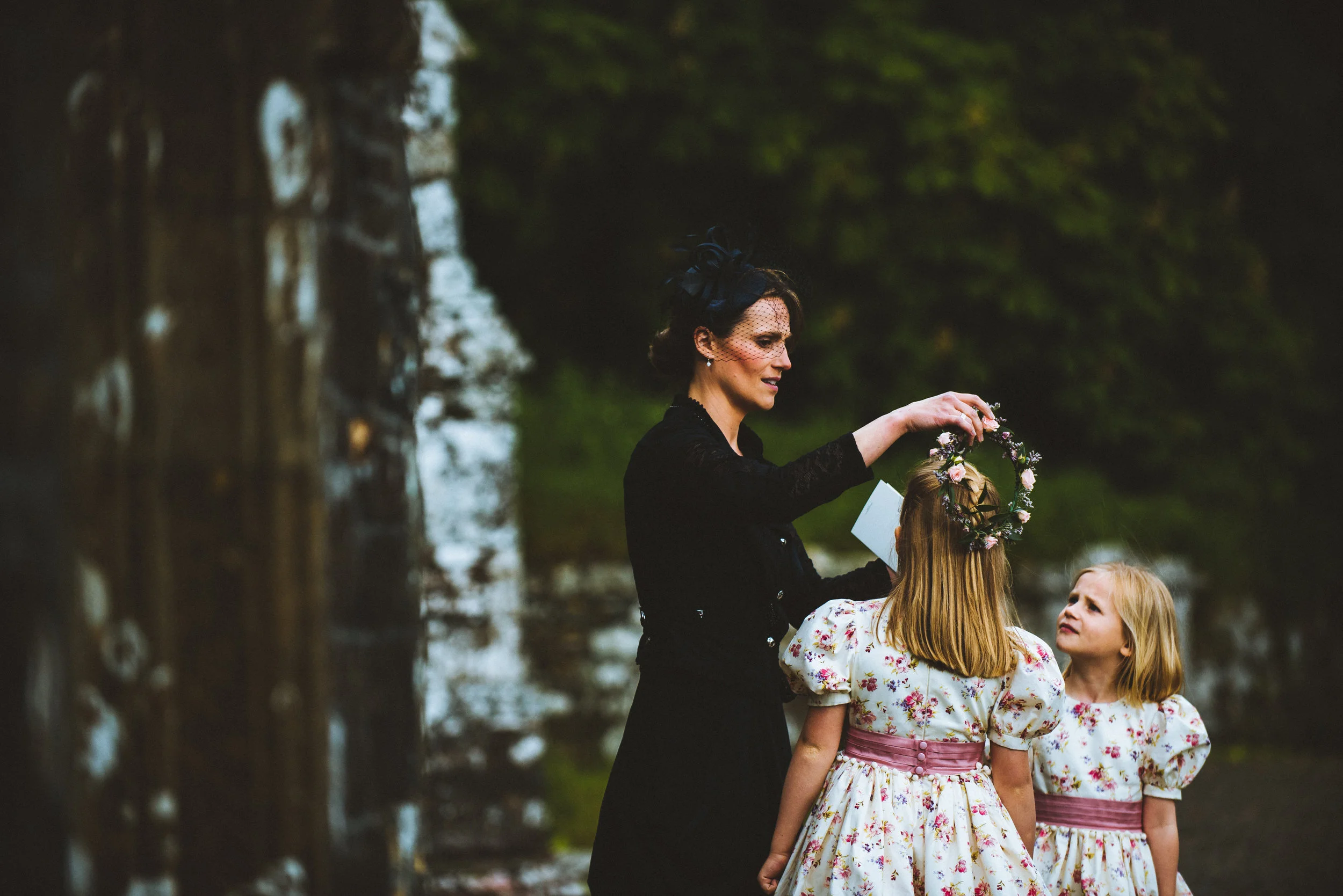 Isles of scilly photography. A woman in black dress placing a floral crown on a girl’s head during an outdoor ceremony, with another girl watching, by a waterfall with lush greenery in the background.