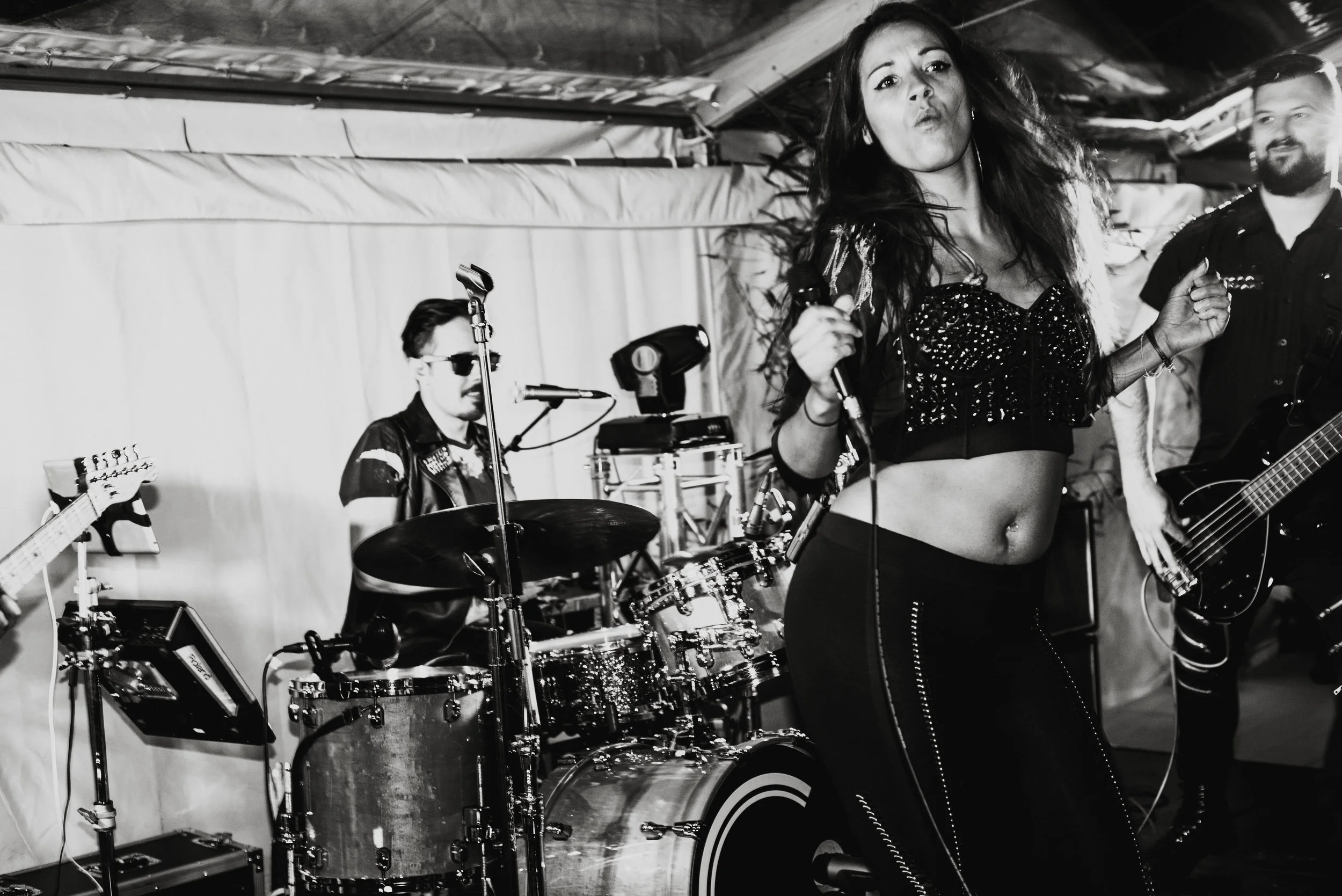 A woman singing into a microphone on stage with a band behind her, playing instruments including drums, guitar, and bass, in a black and white photo.