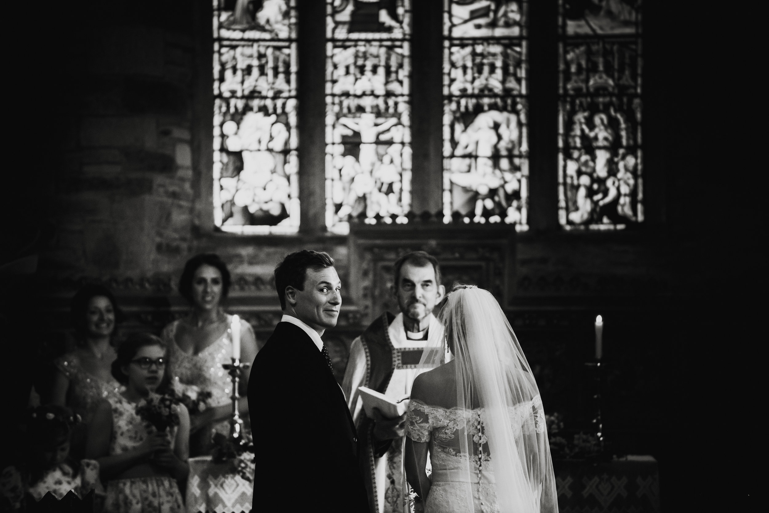 A black and white photo of a wedding ceremony inside a church, showing a bride and groom facing each other with a priest standing behind them, and bridesmaids and children in the background.