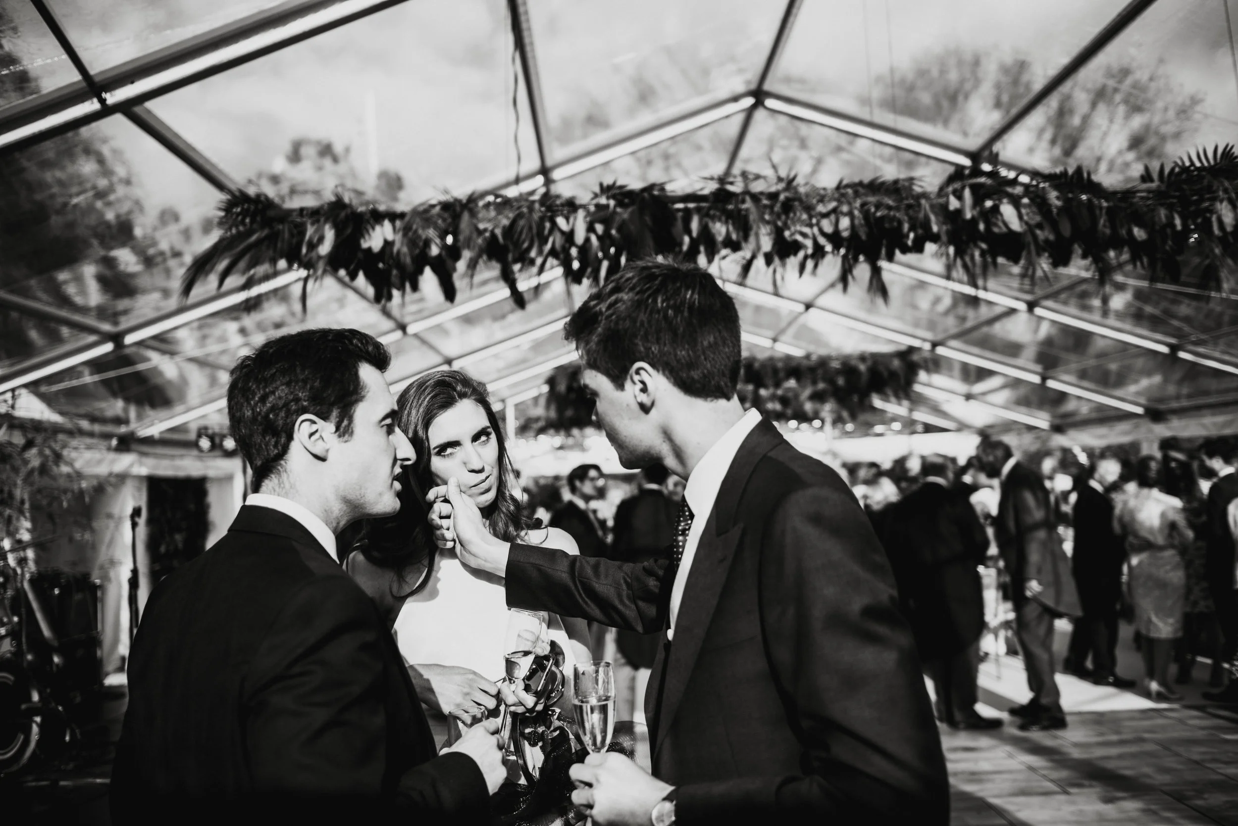 Three people at a social event, two men and a woman, engaging in conversation under a decorated transparent tent ceiling with hanging foliage.