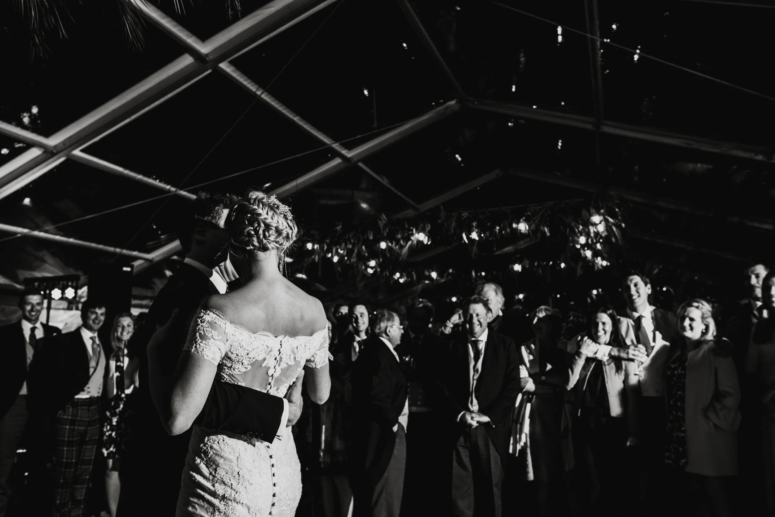 Black and white photo of a wedding reception with bride and groom dancing, surrounded by onlookers in formal attire.