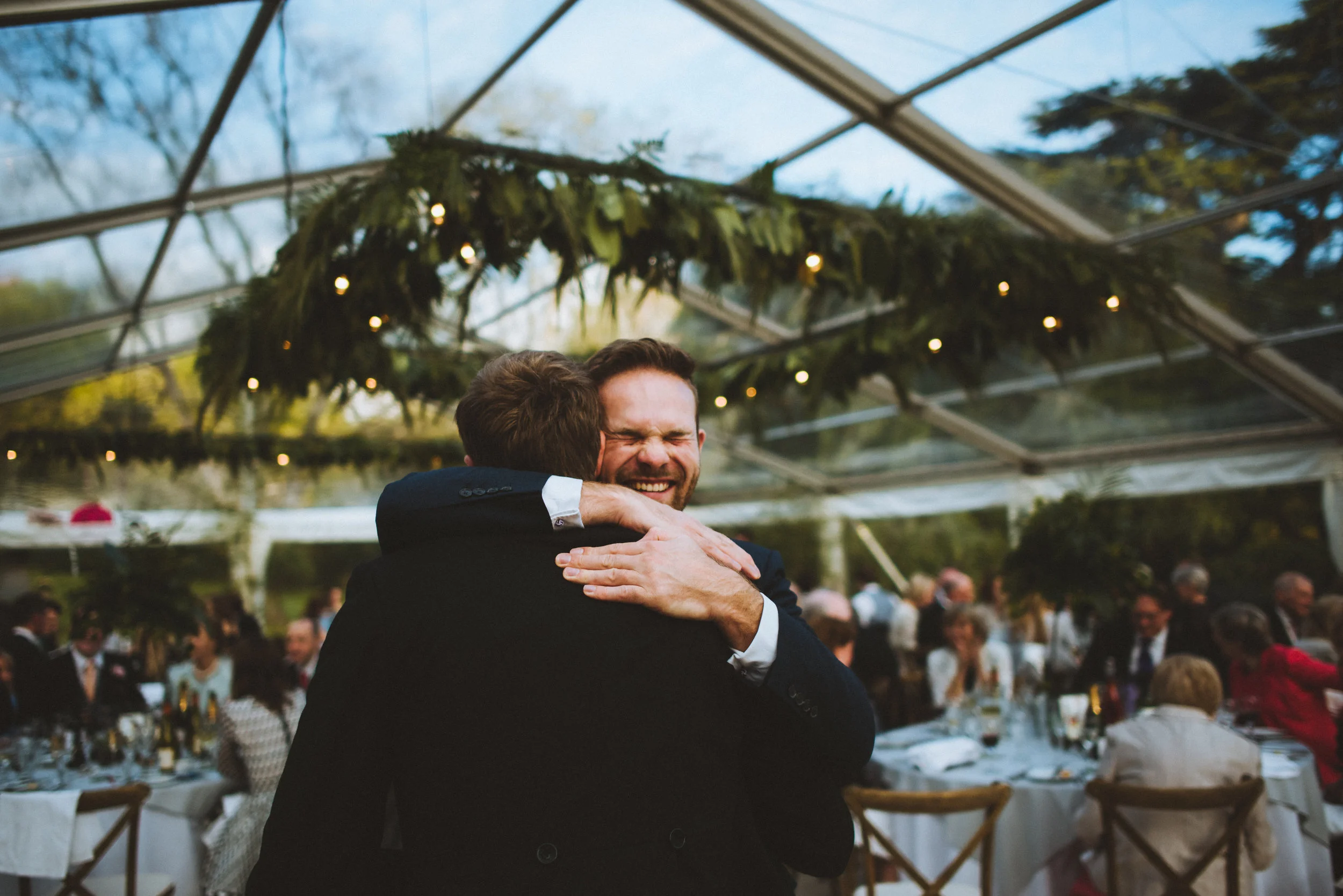 Two men in suits hugging at a wedding reception, with tables and guests in the background under a glass ceiling decorated with greenery and string lights.