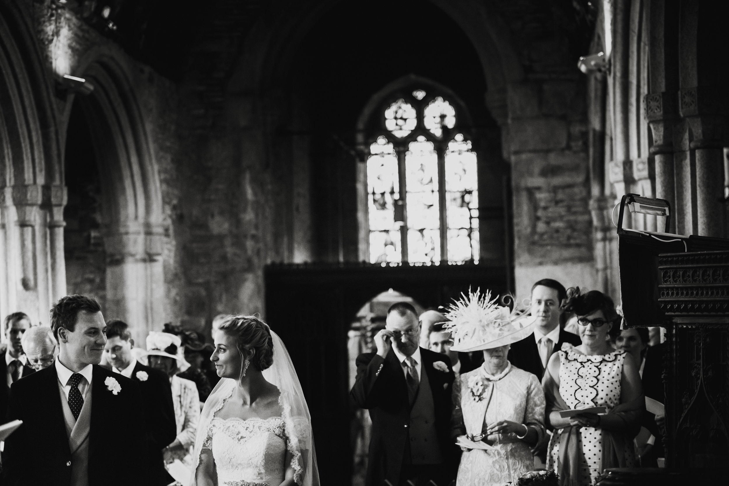 A black and white photo of a wedding ceremony inside a church, with the bride and groom standing together and guests nearby.