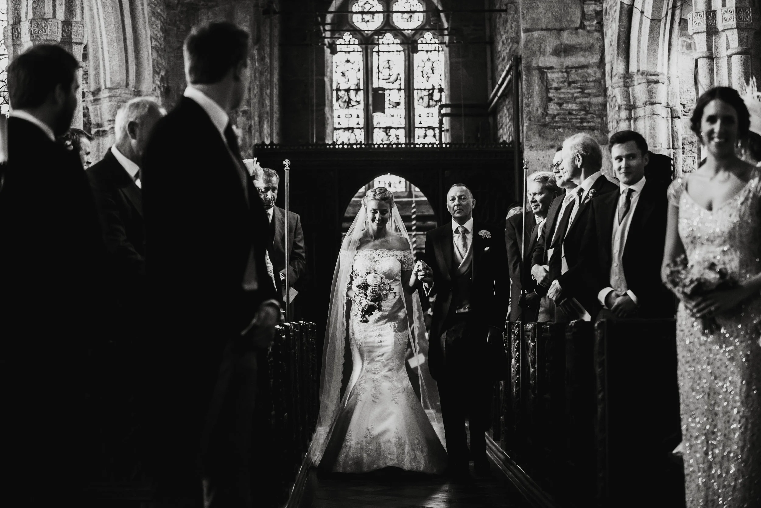 A black and white photo of a bride walking down the aisle with her father in a church, surrounded by guests - Captured by Cornwall based wedding photographer Mark Shaw