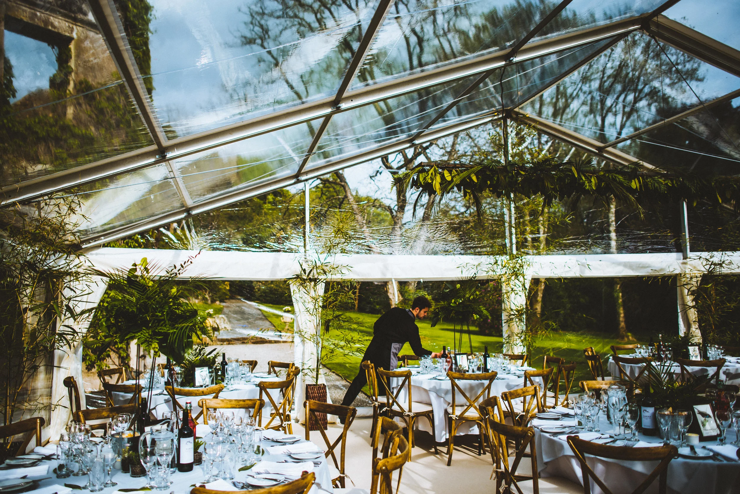 A man arranging tables in an event space with glass walls and ceiling, surrounded by greenery.