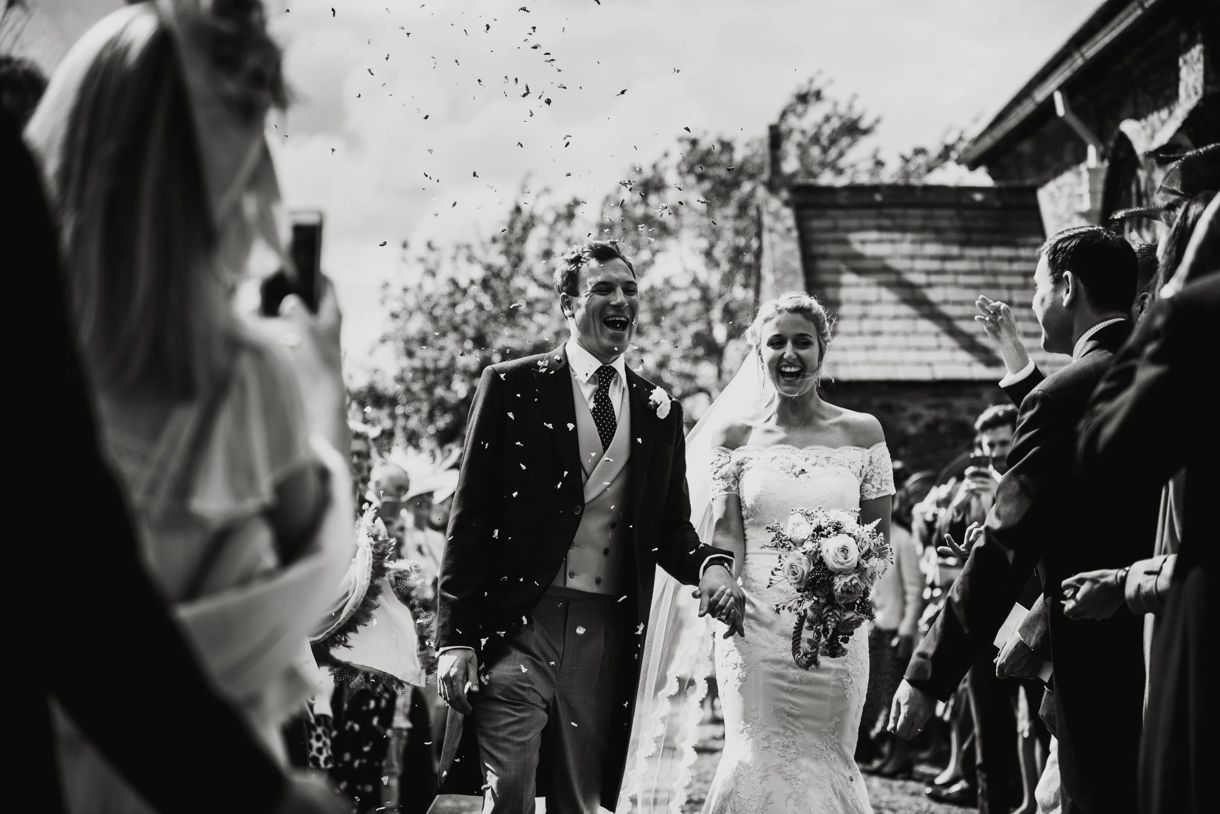 Wedding photo of A black and white photo of a happy bride and groom walking hand in hand surrounded by guests at an outdoor wedding, with confetti in the air.
