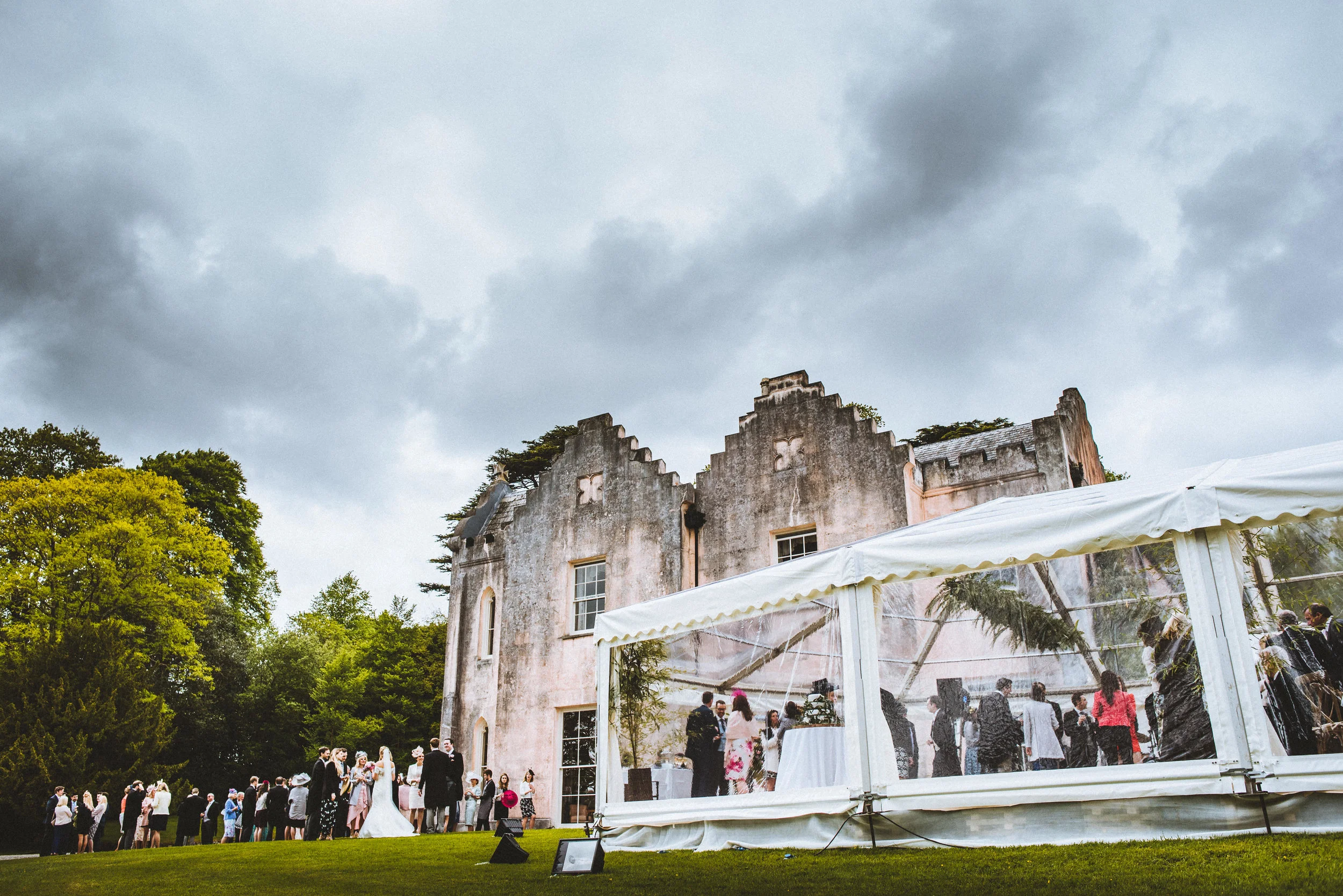 Guests at an outdoor wedding reception under a tent, with a historic castle-like building in the background on a cloudy day.