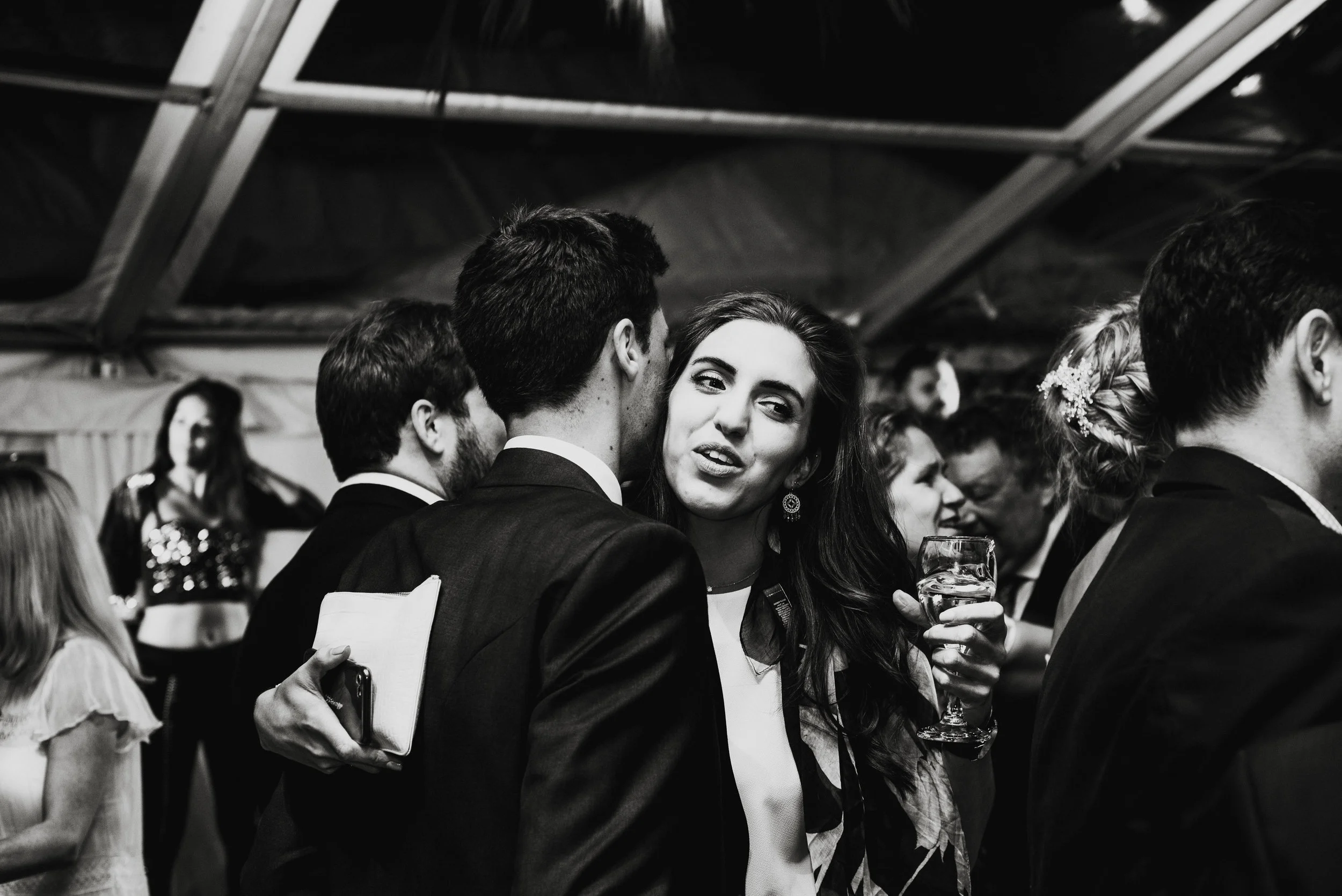 A black and white photograph of a social gathering, showing a woman with long dark hair holding a glass of wine and talking to a man in a suit. There are several other people in the background engaged in conversations.