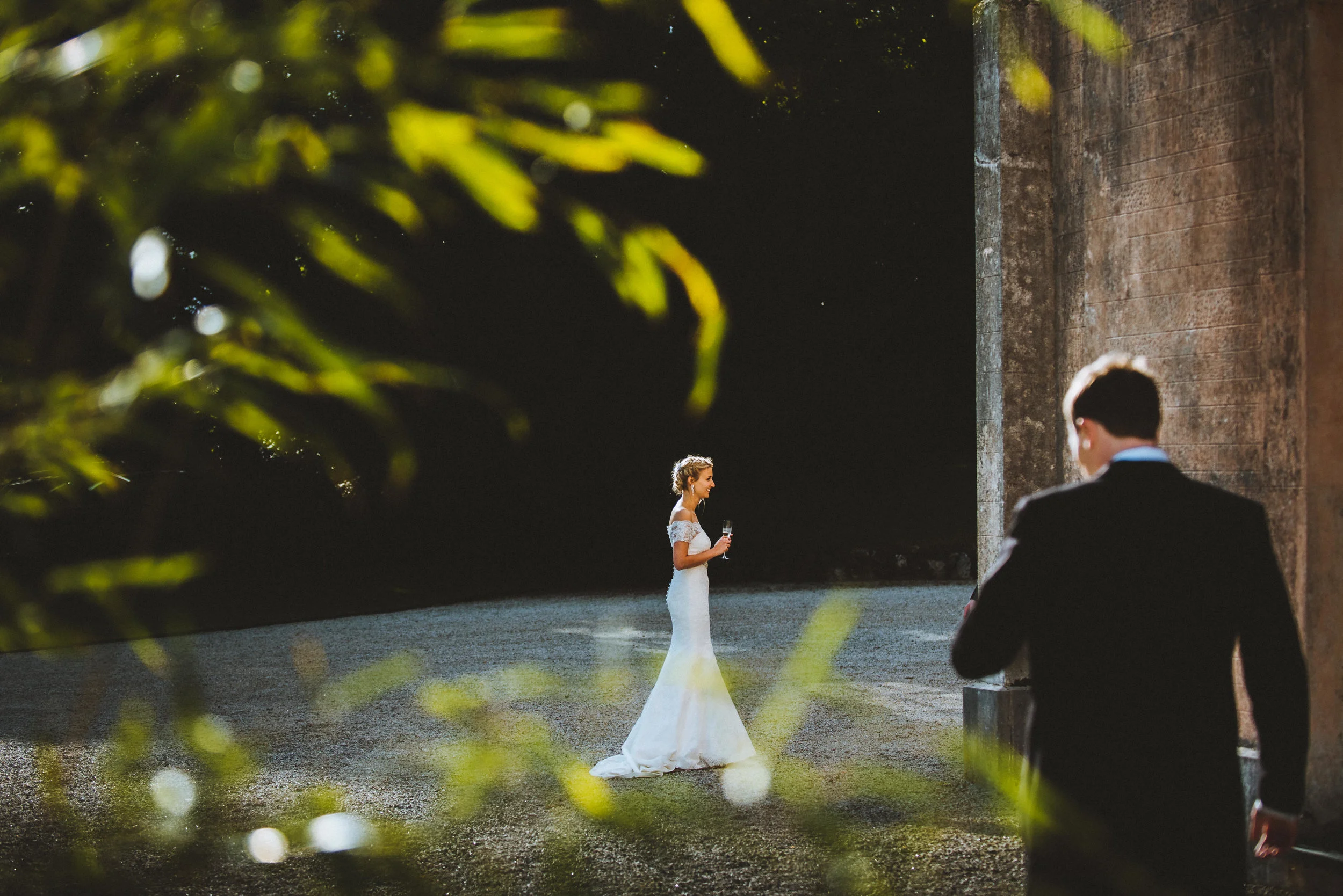 A bride in a white wedding dress holding a glass of champagne, standing outdoors near a brick building with a man in a black suit approaching her, framed by blurred green leaves in the foreground.