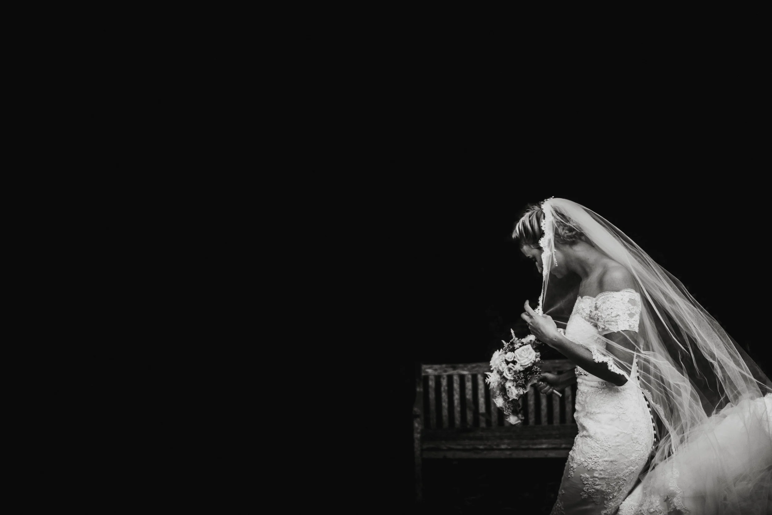 Isles of scilly photography. A bride in a lace wedding dress with a veil holding a bouquet, standing on a bench, looking down and appearing emotional.