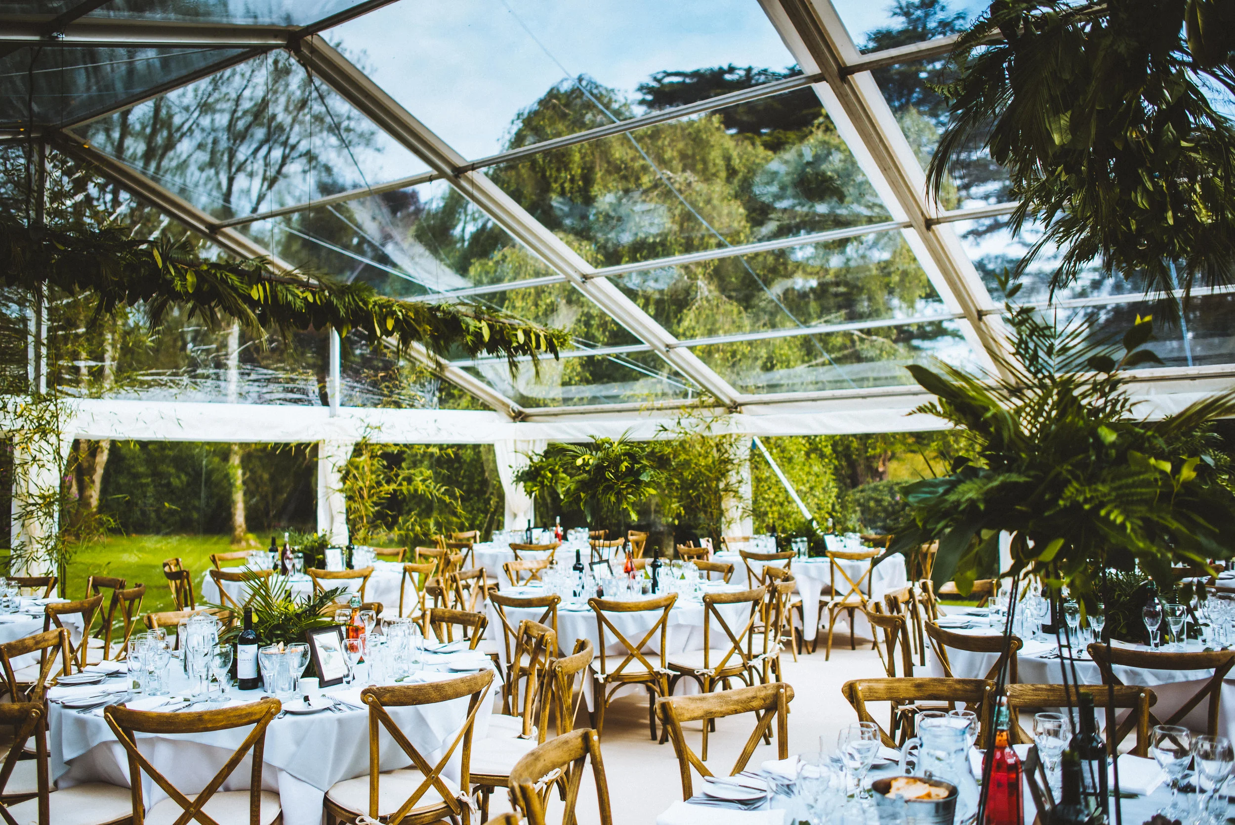Indoor dining area with white tablecloths, wine bottles, and glassware, surrounded by wooden chairs, inside a transparent tent with a view of green trees and blue sky.