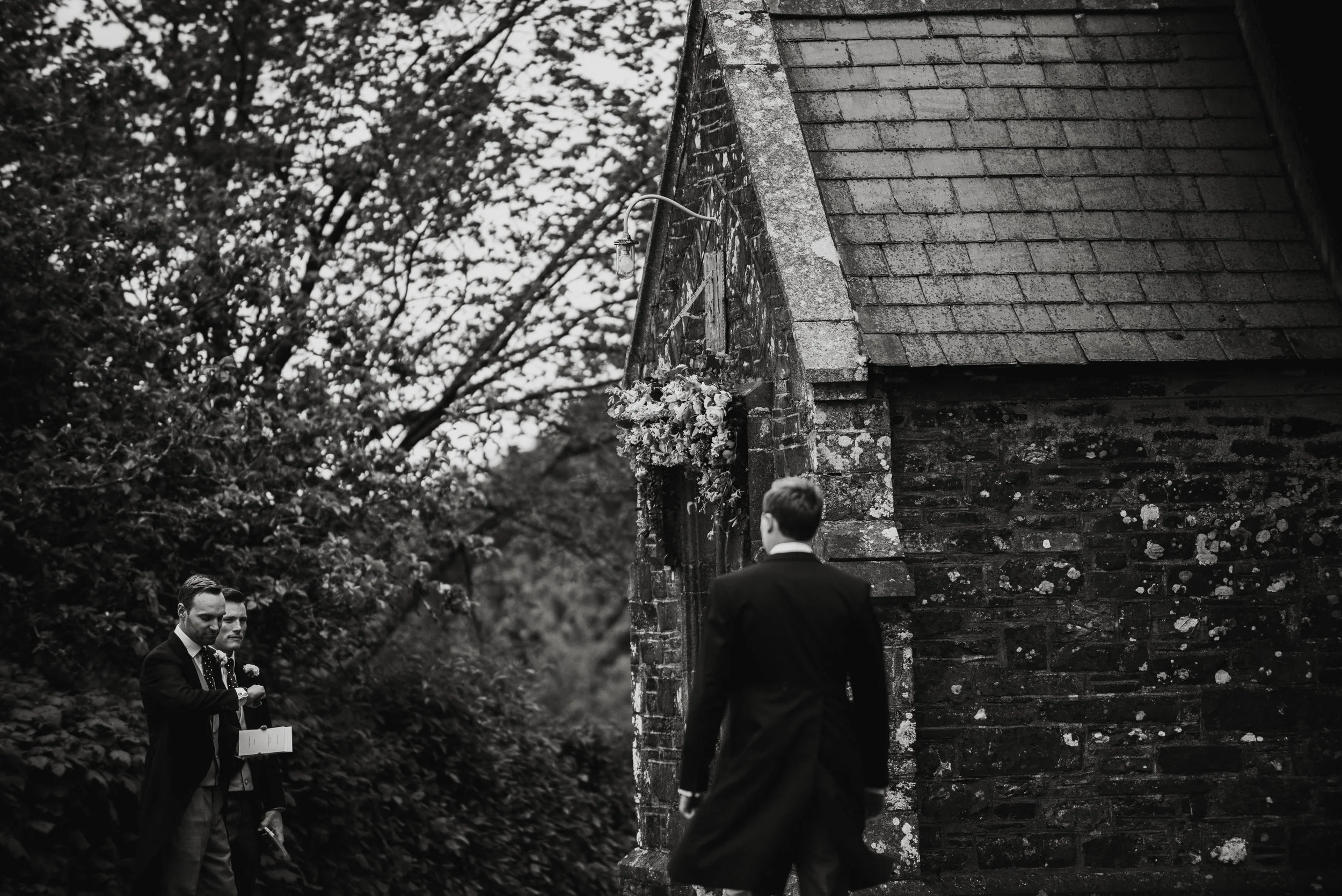 Isles of scilly photography. Three men in formal suits outside a stone church, with one man walking toward the building while two others stand nearby holding a book or a phone, in a black and white photo.