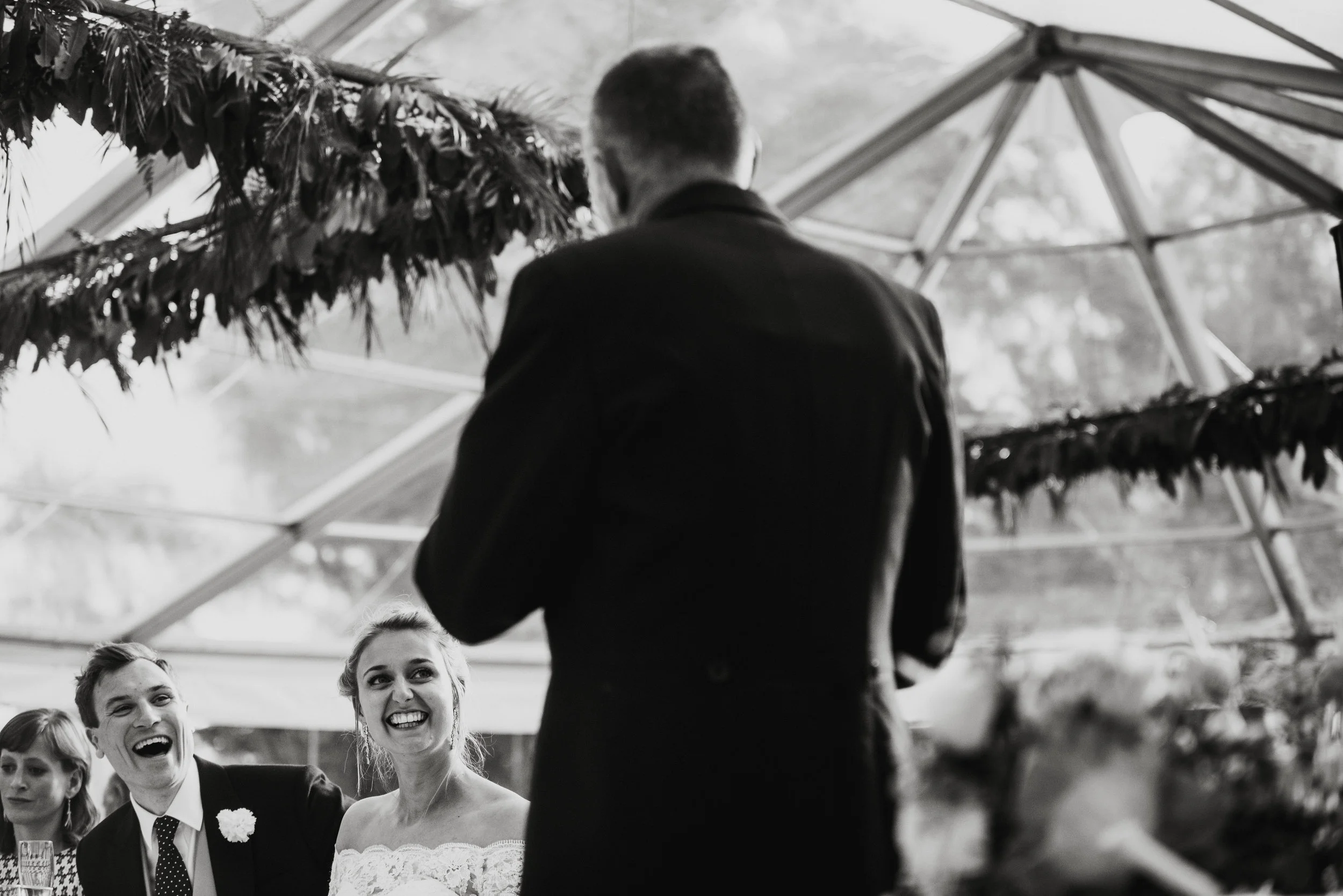 Black and white photo of a wedding reception showing a bride and groom smiling and laughing at a speech, with a person in a suit speaking in front of them, inside a glass tent with foliage and decorations.