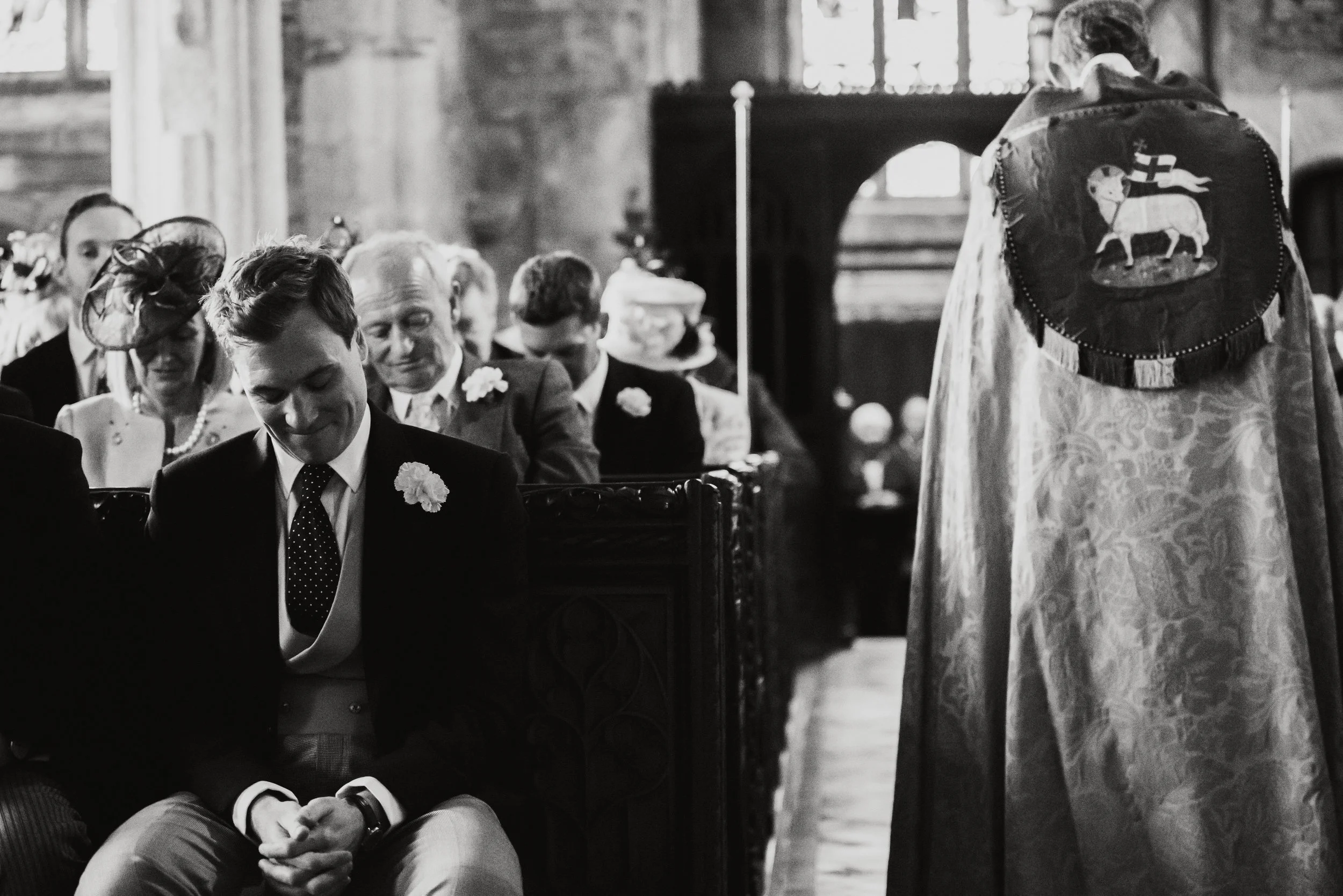 Black and white photo of a wedding ceremony inside a church, with several seated guests and a priest standing on the right side.