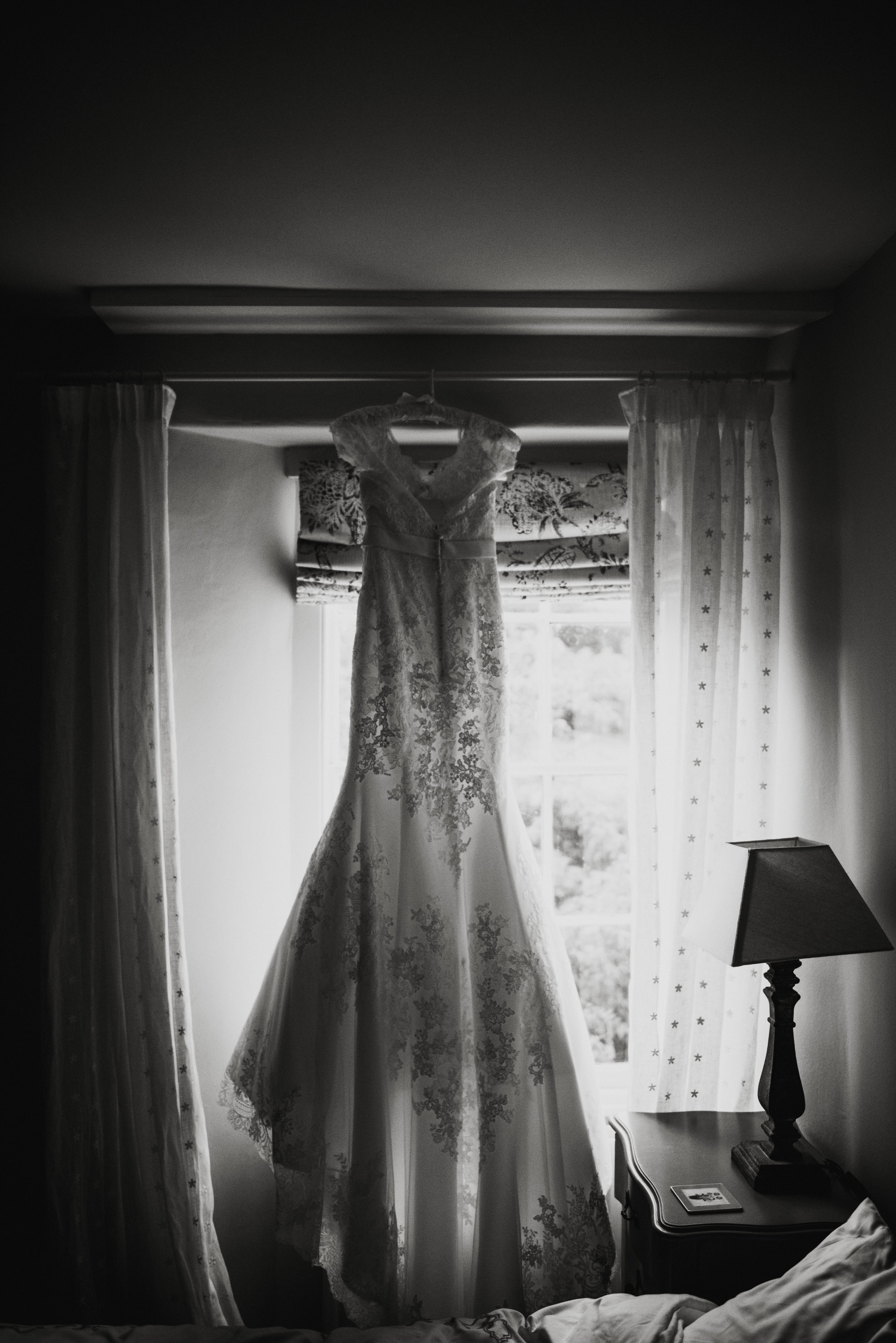 A wedding dress hanging in front of a window in a bedroom, with curtains on either side and a bedside table with a lamp and a framed photo.