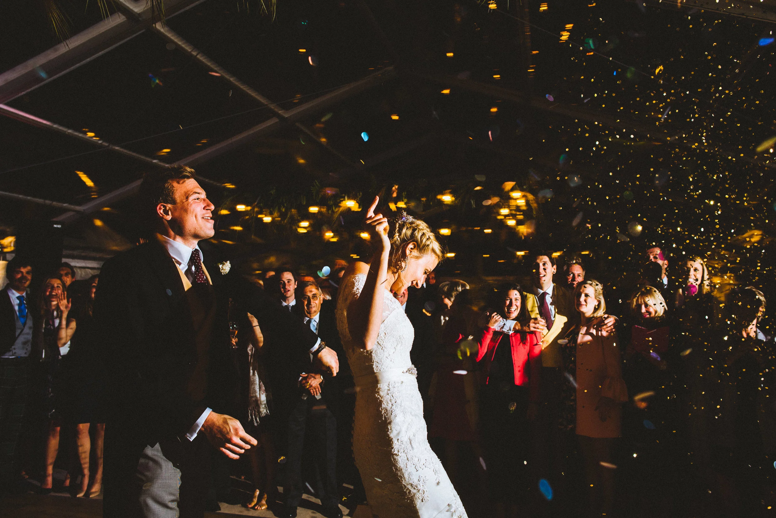 A bride and groom dancing at their wedding reception with guests celebrating around them and golden confetti falling from above.