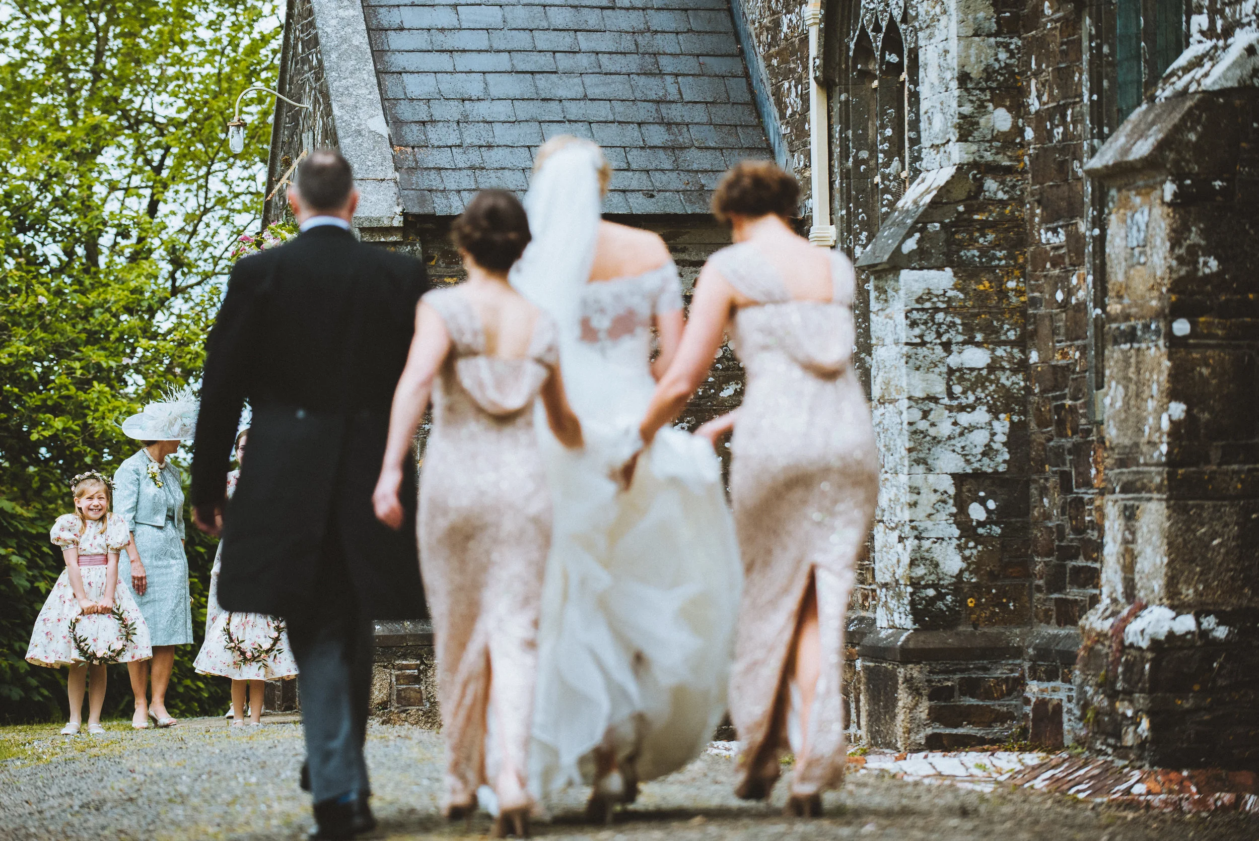 A wedding scene with three women, a man, and two young girls outside an old stone building. The women are in elegant dresses, and the man is in a tuxedo. The young girls are in floral dresses, one holding a small wreath, smiling and observing the sce