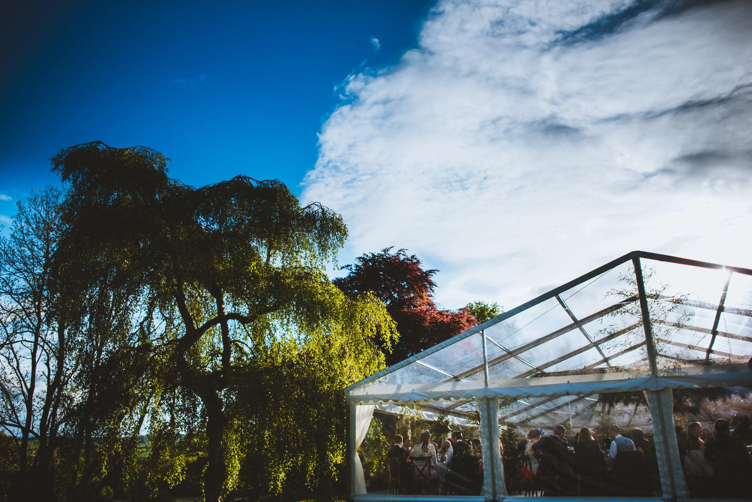 Outdoor event with people dining inside a transparent tent, surrounded by green trees under a partly cloudy sky.