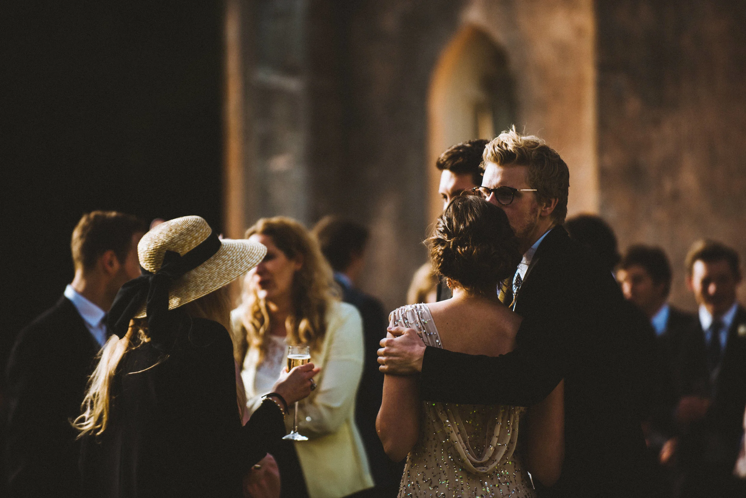 A couple hugging at a formal event, with other guests socializing in the background - Captured by Cornwall based wedding photographer Mark Shaw