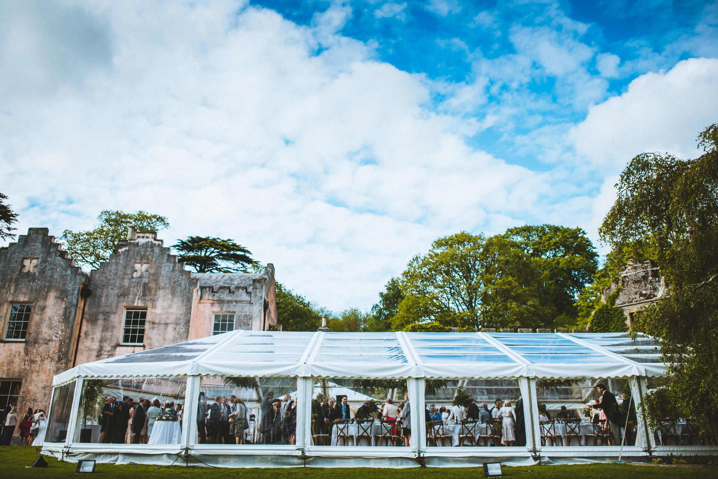 Outdoor event taking place inside a large white tent set up on a grassy area near historic buildings, with people gathered inside and some mingling outside, under a partly cloudy sky.