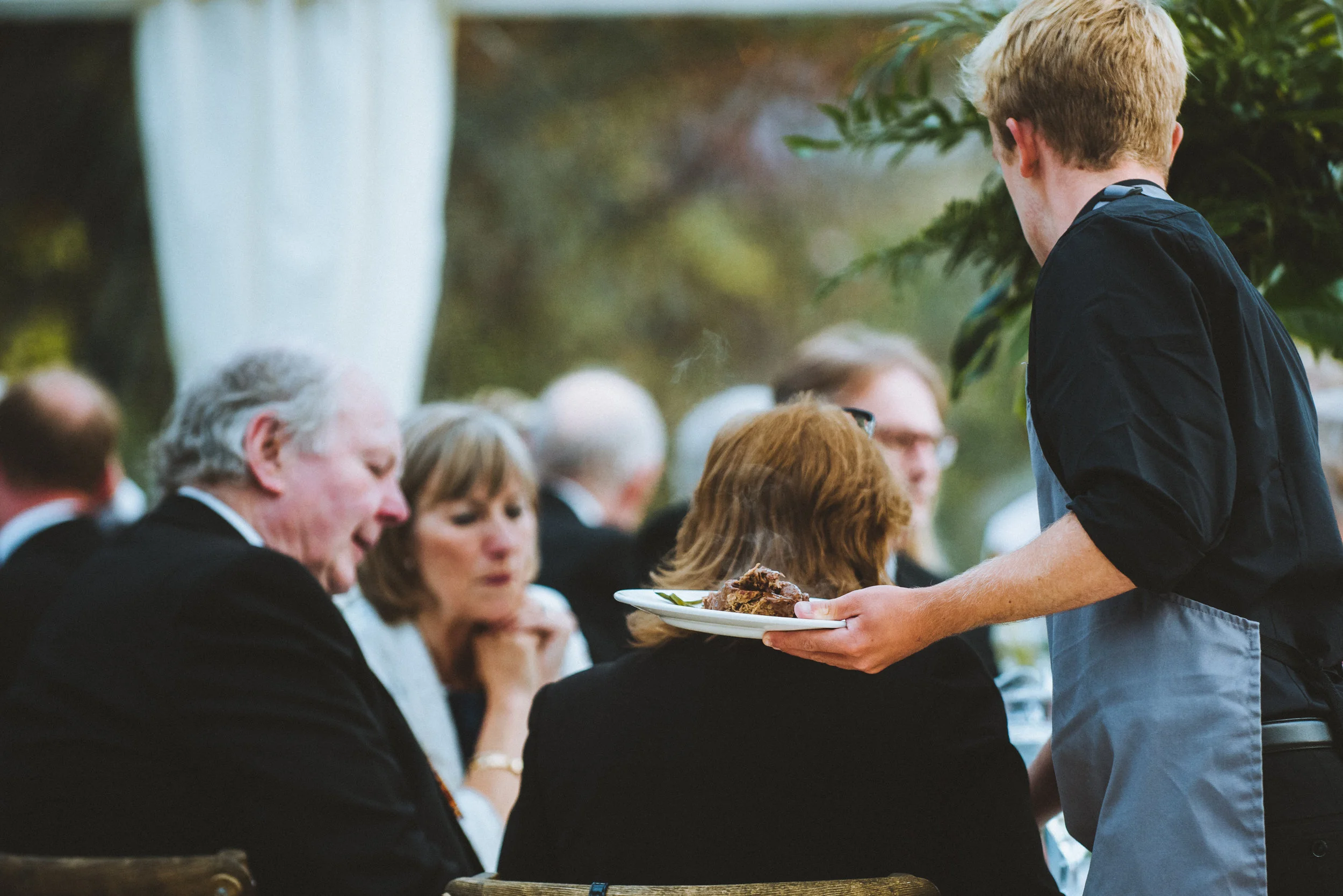 A waiter serves food to a woman seated outdoors at a formal gathering, with other guests sitting nearby, during the daytime.