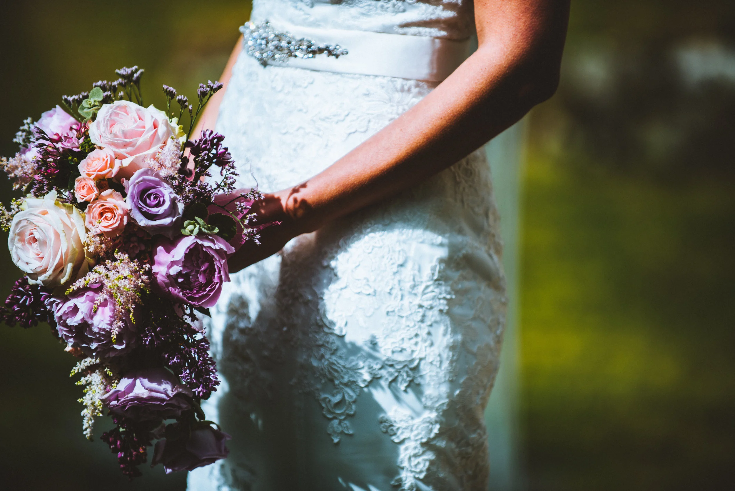 Isles of scilly photography. Close-up of a bride in a white lace wedding dress holding a bouquet of pink, purple, and white roses with greenery, outdoors with blurred green background.