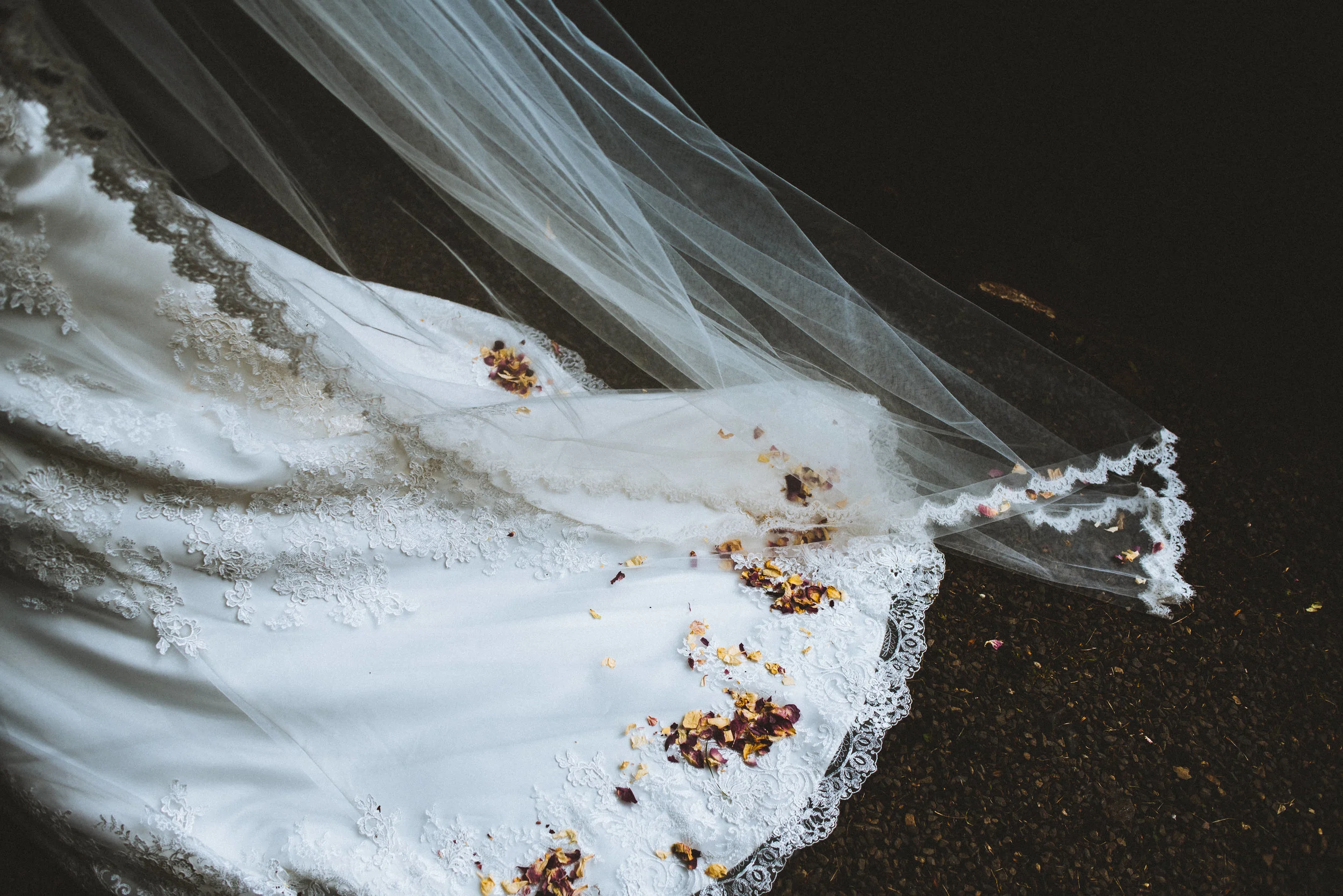Wedding photo of wedding dress with lace details and a veil, draped on the ground with scattered flower petals, photographed from above on a dark surface.