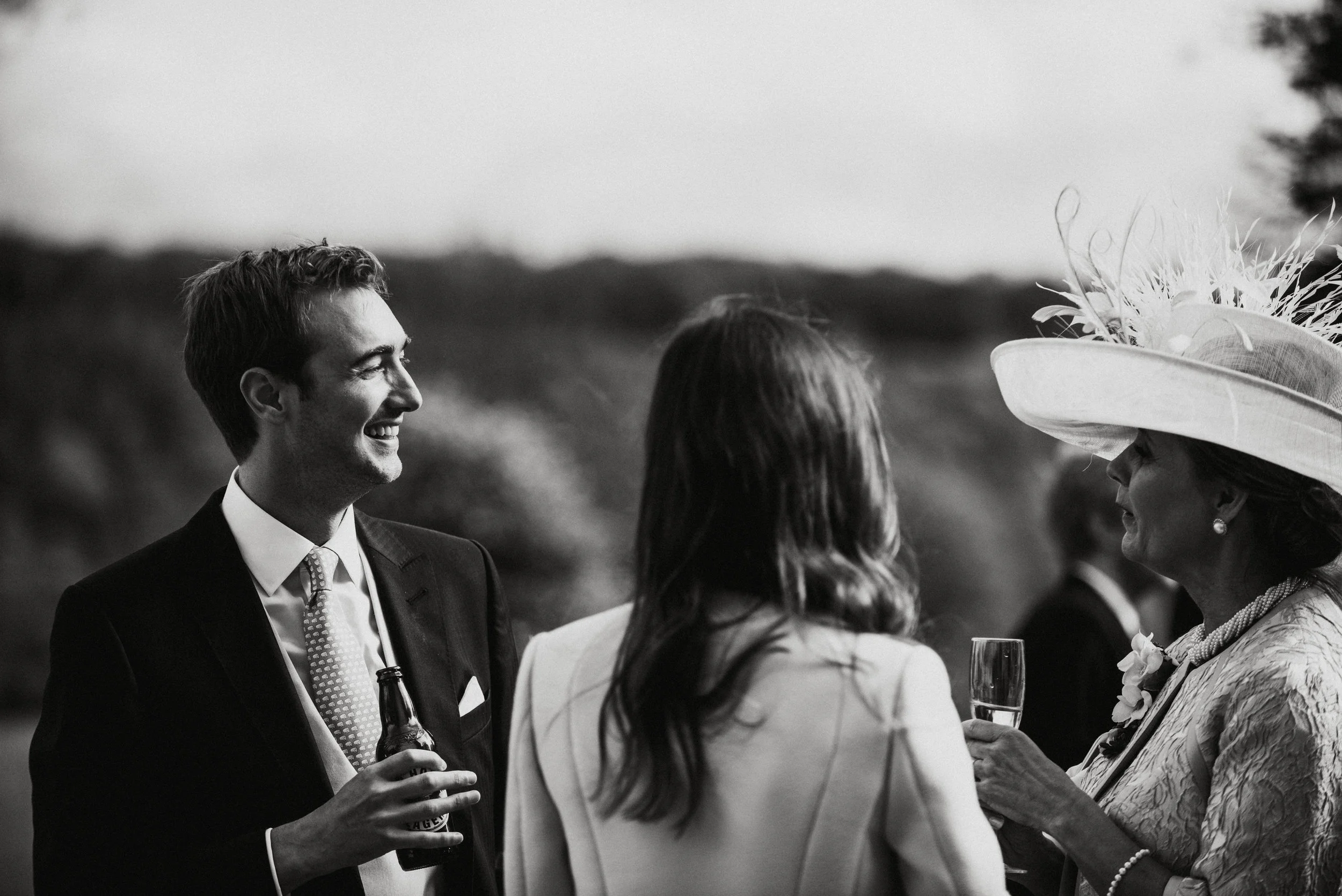 Black and white photo of a wedding reception with a man in a suit holding a beer bottle, a woman with long hair, and an older woman in a fancy hat holding a glass of champagne, engaged in conversation outdoors.