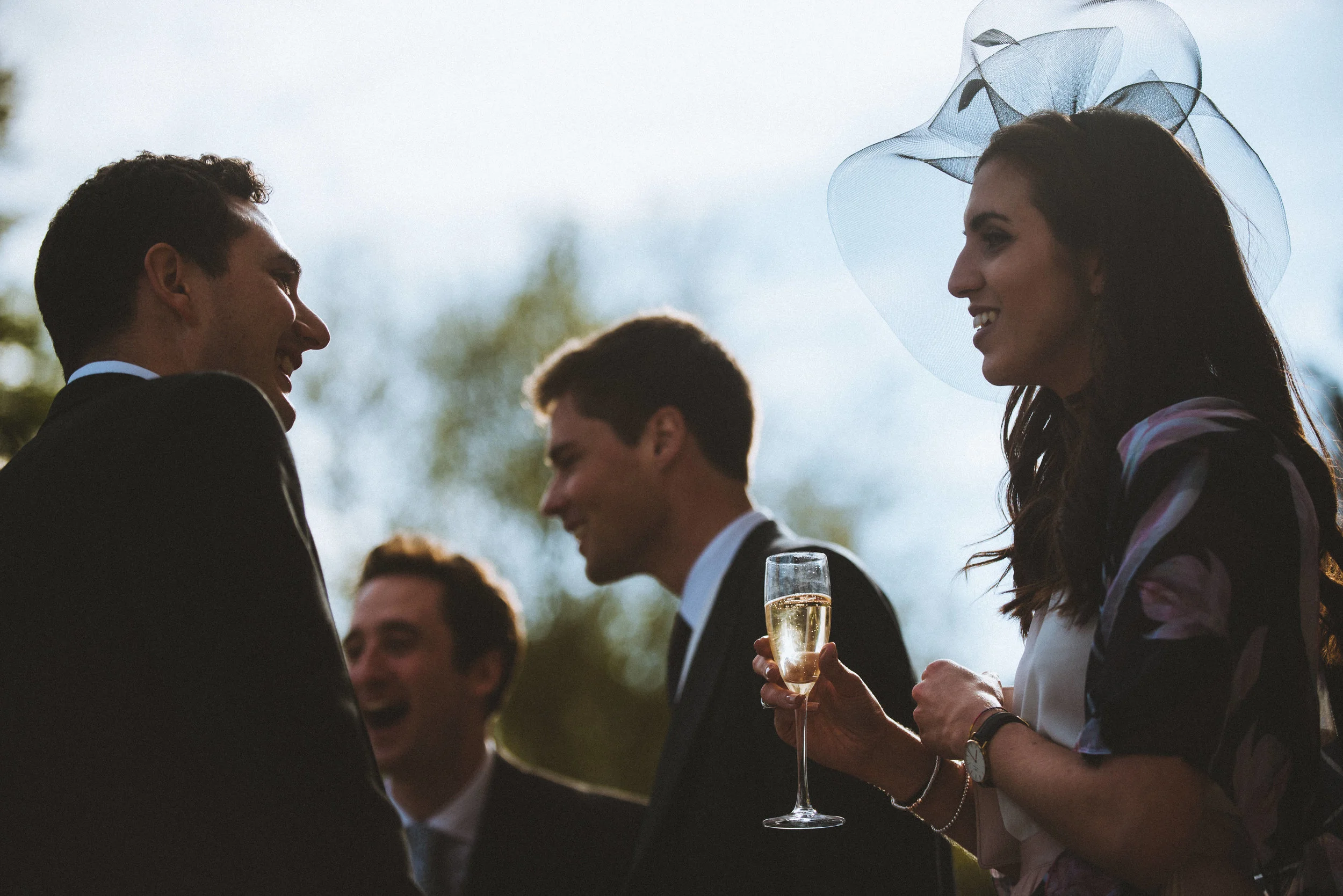 Four people dressed in formal attire, holding glasses of champagne, engaging in conversation outdoors during daytime, with blurred trees in the background.