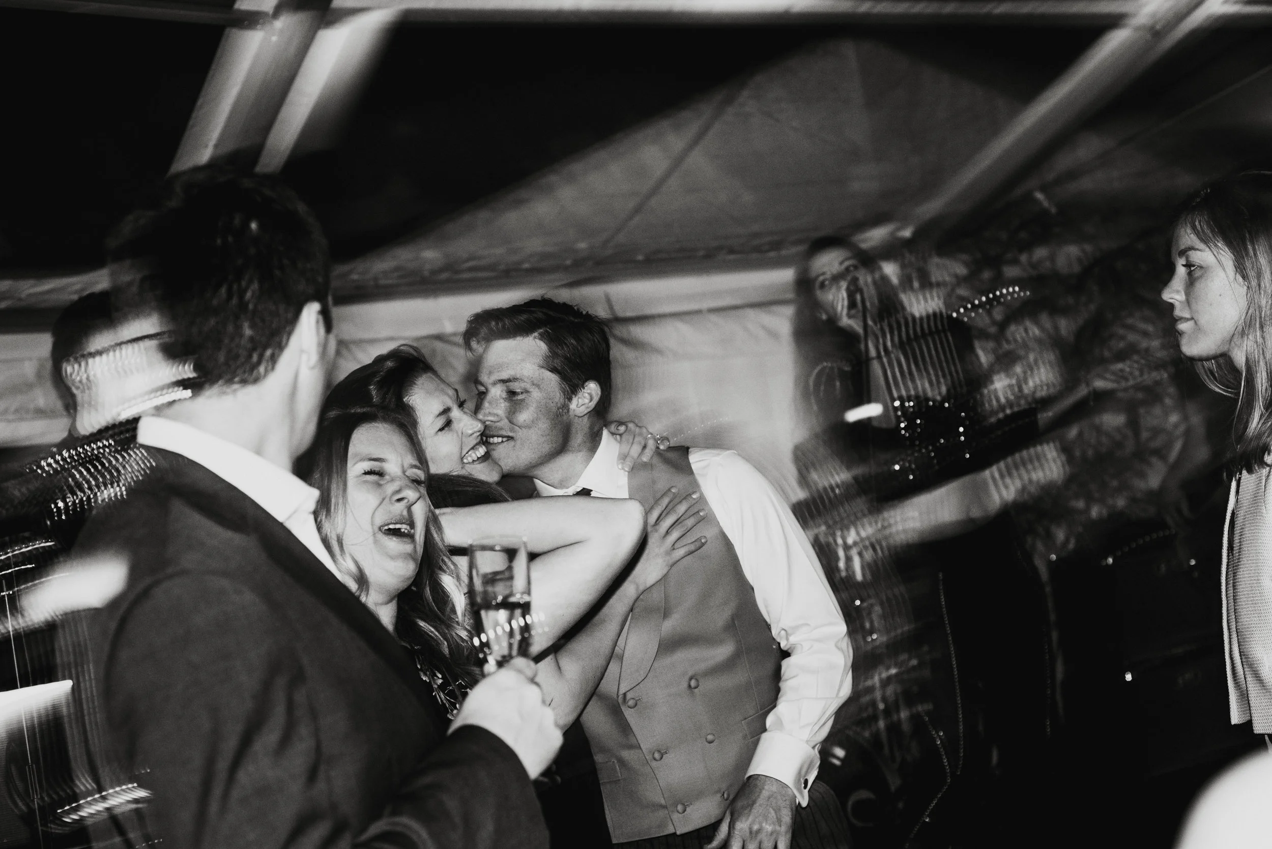 Four people at a celebration, with one person holding a glass of champagne, a woman hugging a smiling man, and others looking on.