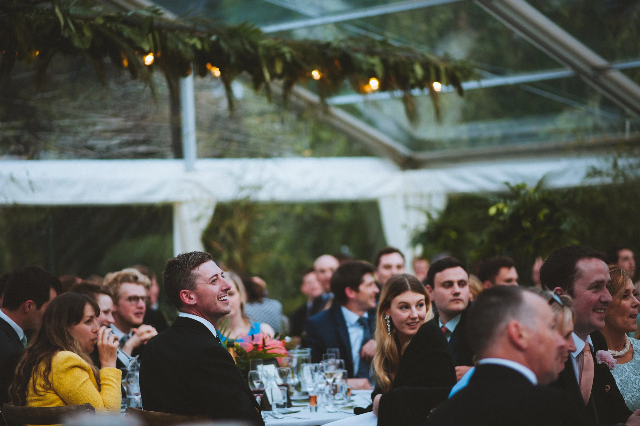 Guests sitting at a banquet table inside a glass greenhouse or tent, smiling and listening to a speaker at an event or celebration.