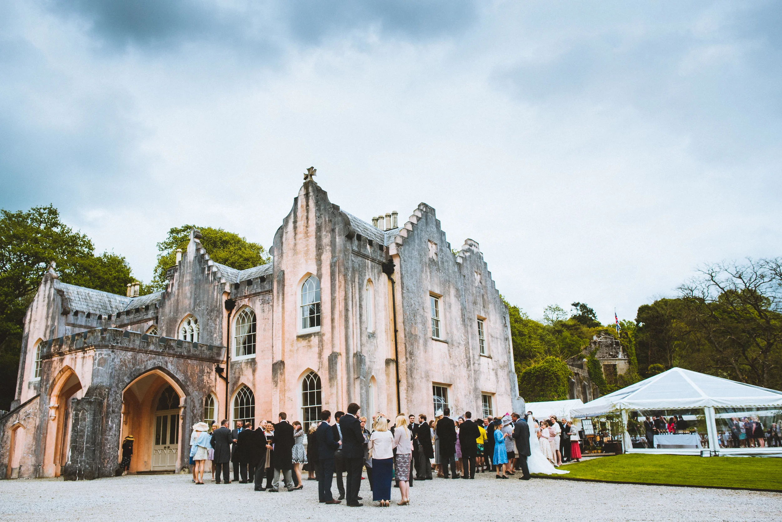 A group of people gathered outside an old, castle-like building with gray, weathered walls, arched windows, and a steeply pitched roof, with trees and a sky in the background during daytime.