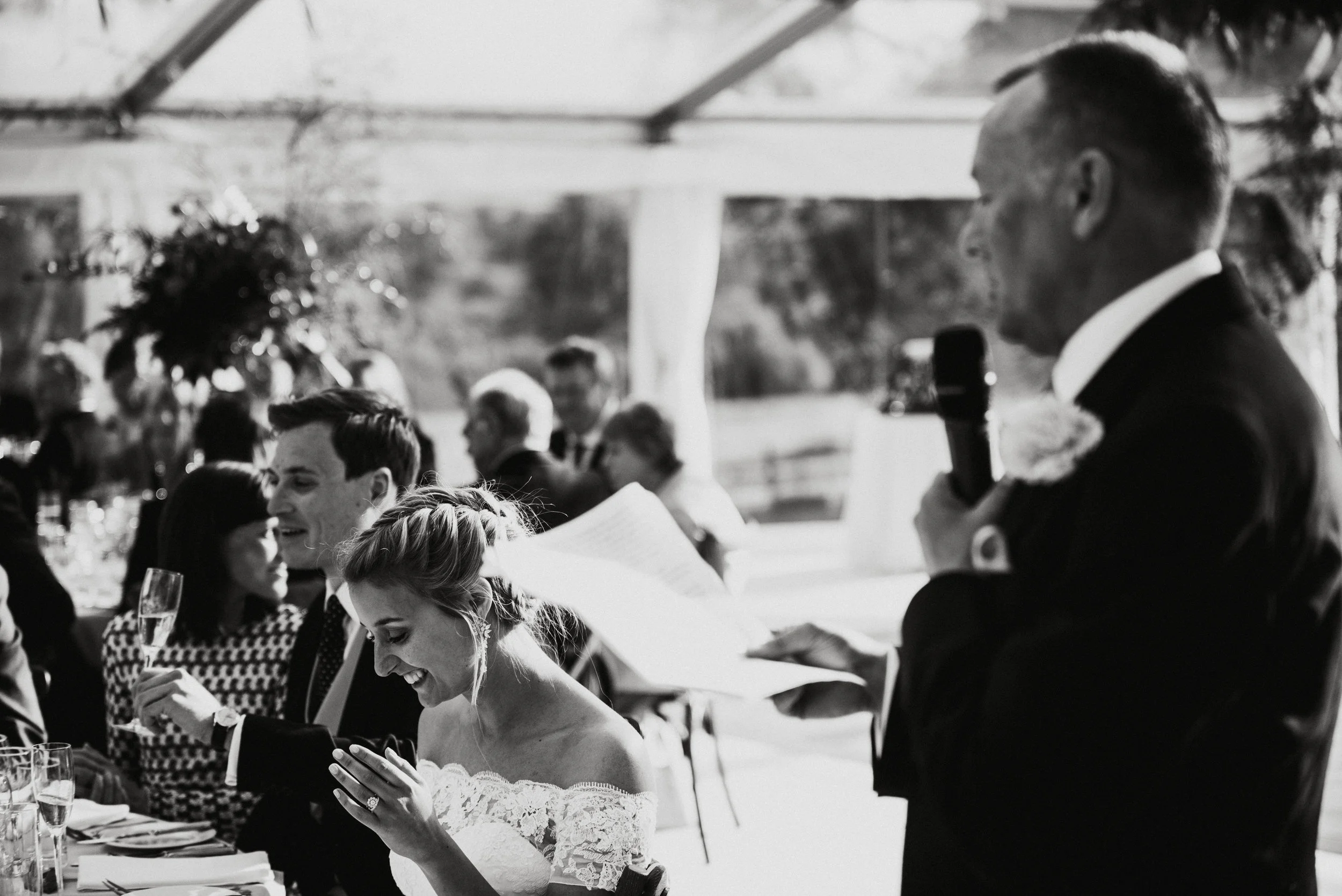 A wedding reception with guests seated at a table, some laughing and smiling, with a woman in an off-the-shoulder lace dress looking at her phone. A man in a tuxedo is giving a speech using a microphone, in an outdoor venue with sunlight and trees in