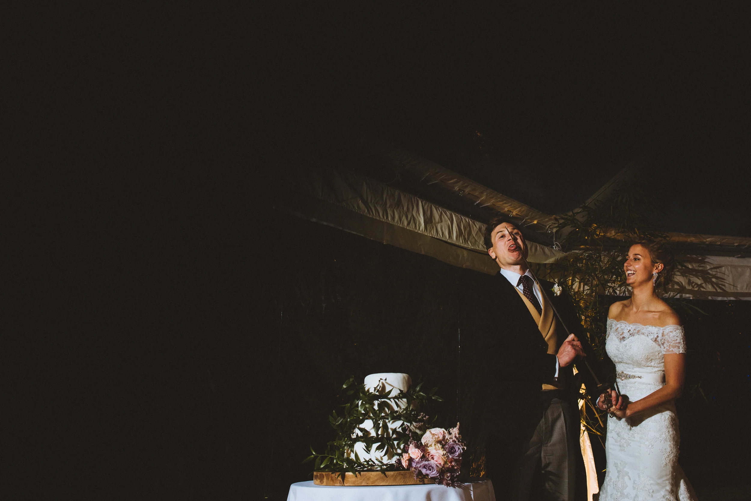 A bride and groom standing together during their wedding celebration, with a wedding cake and floral arrangement on a table in front of them.