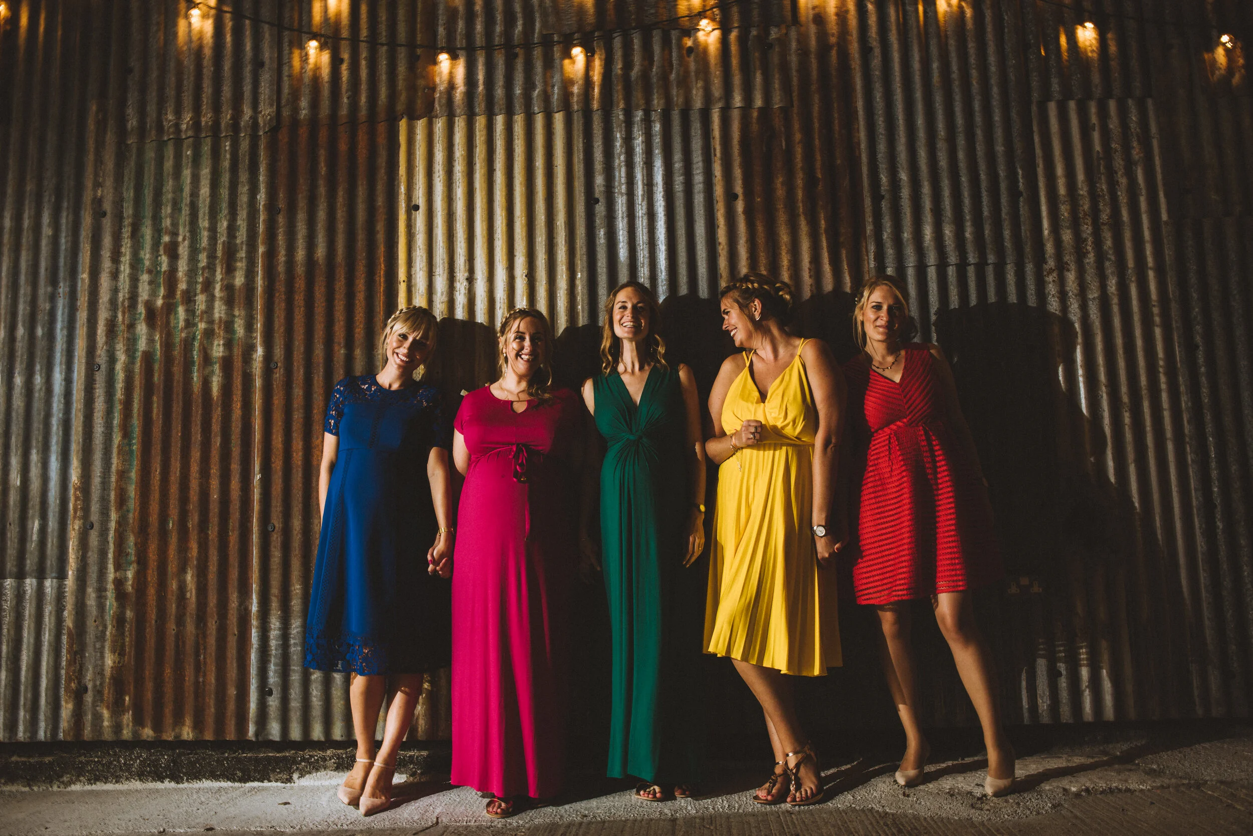 Five women in colorful dresses standing together in front of a corrugated metal wall, smiling and holding hands.