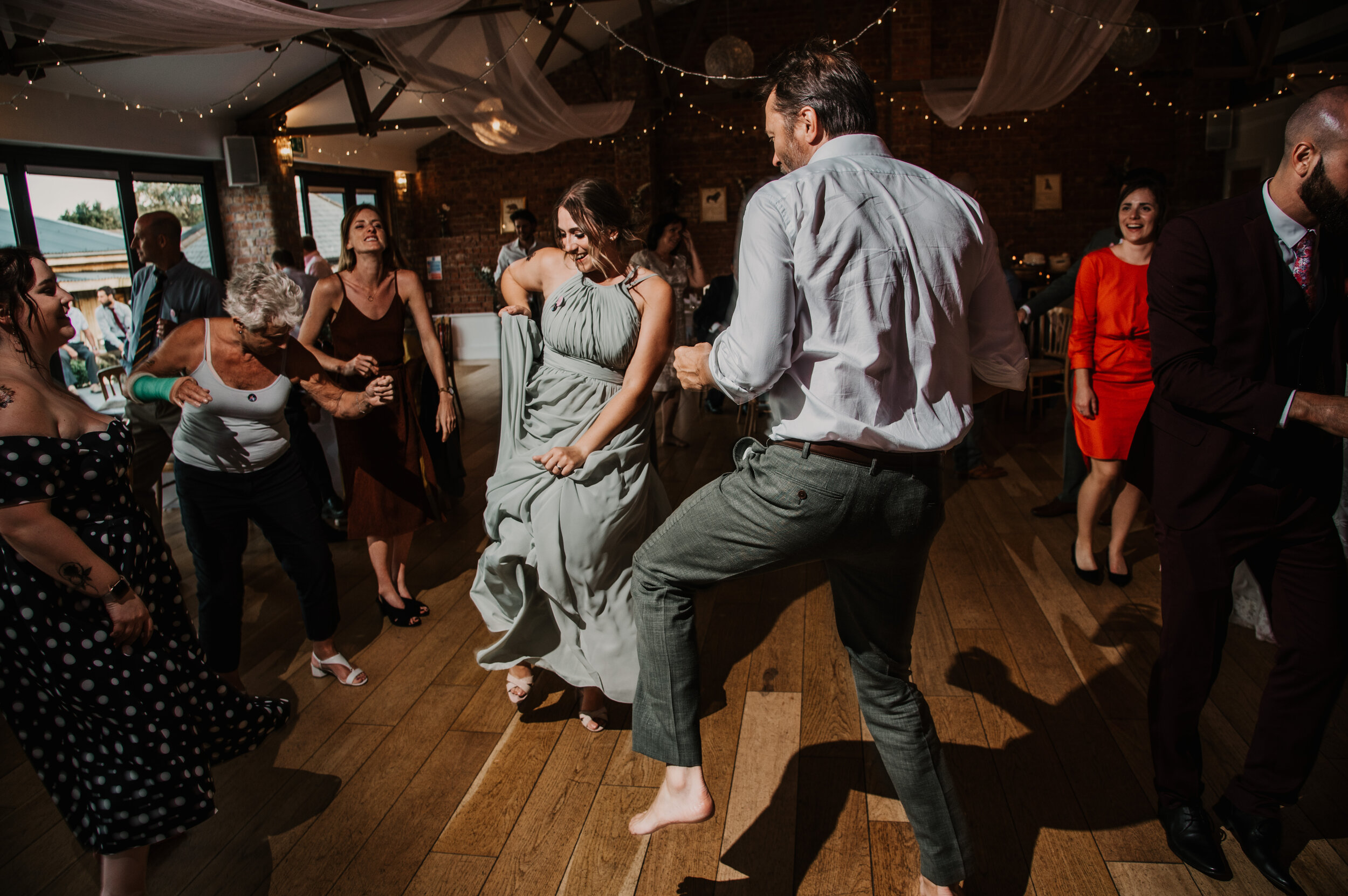 People dancing at a lively celebration indoors with string lights and brick walls.
