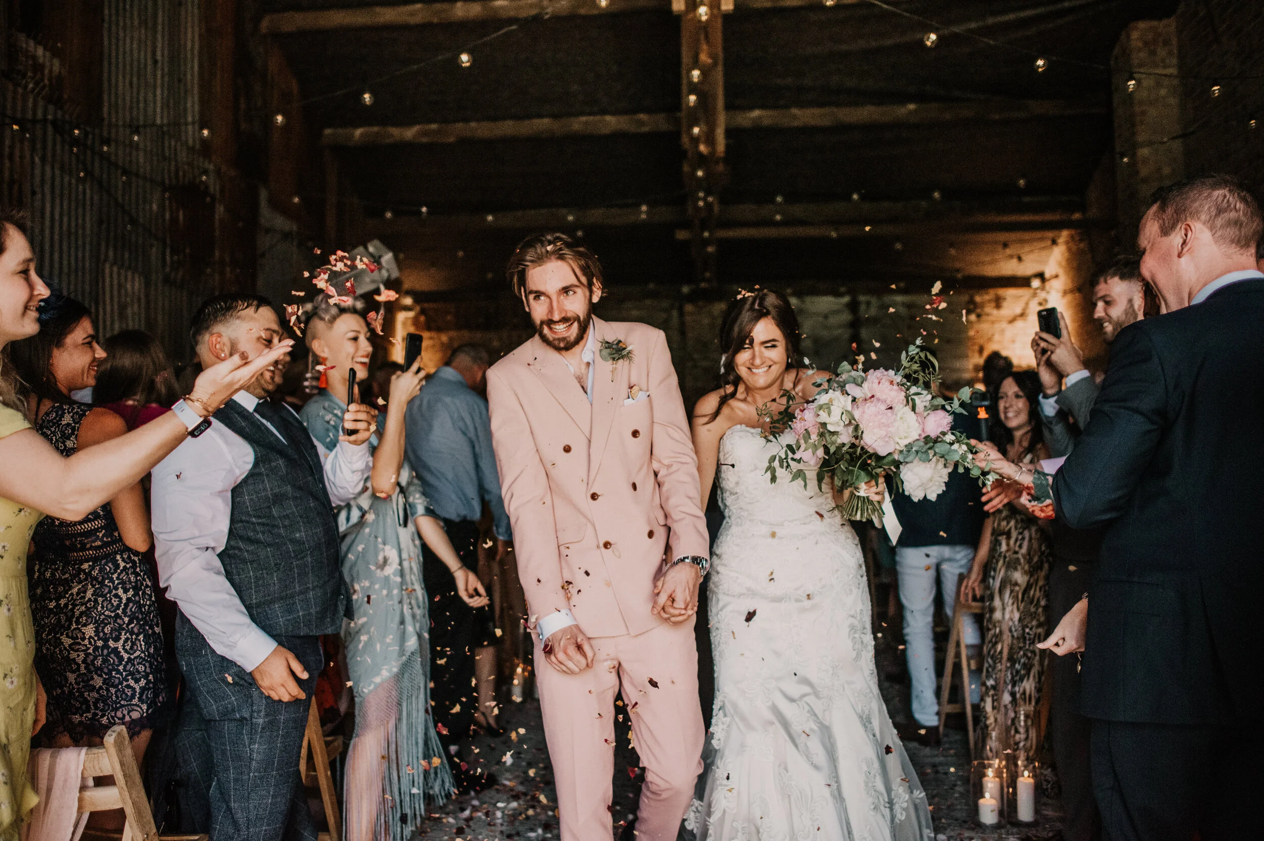 Bride and groom walking down the aisle after a wedding, surrounded by guests throwing confetti and celebrating in a rustic indoor venue.