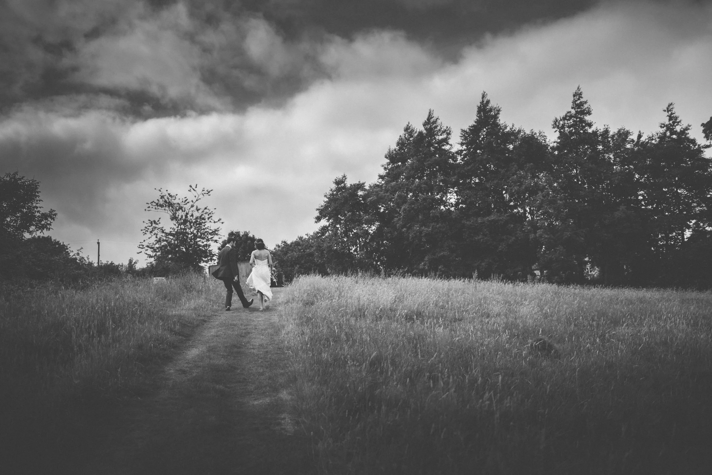 A black and white photograph of a couple walking along a grassy path in a field, with trees and dark clouds in the background.