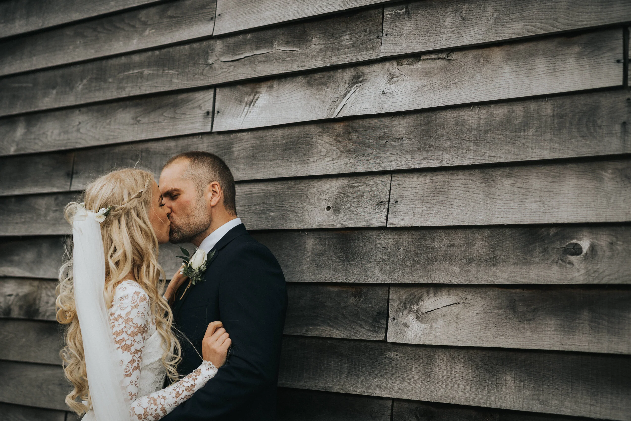 A bride and groom kissing in front of a wooden plank wall, with the bride wearing a long sleeve lace wedding dress and veil, and the groom in a dark suit with a boutonniere.